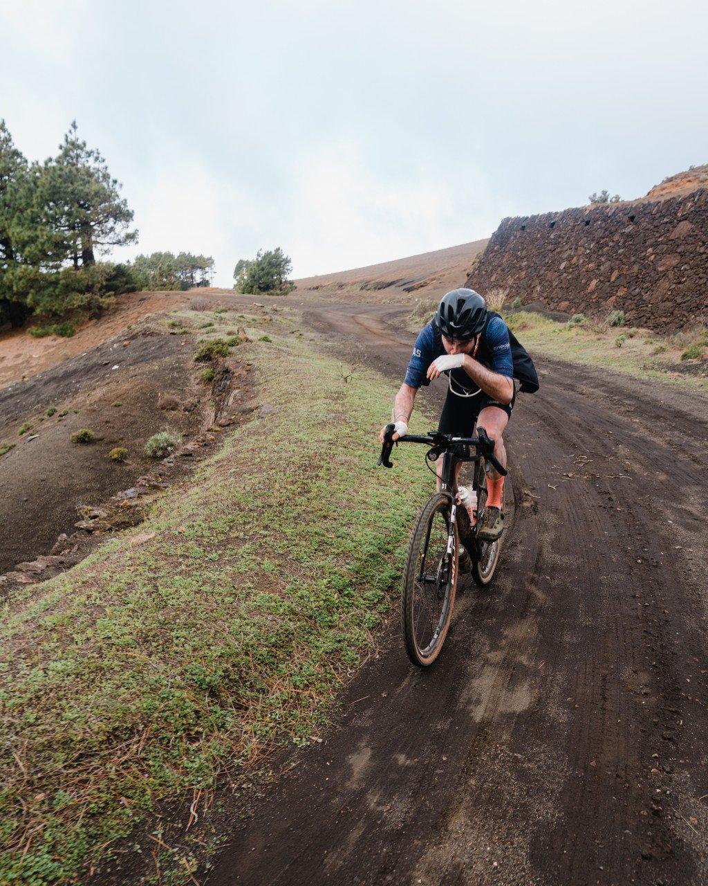 Model wearing blue cycling jersey time trialing on dirt road through hilly terrain