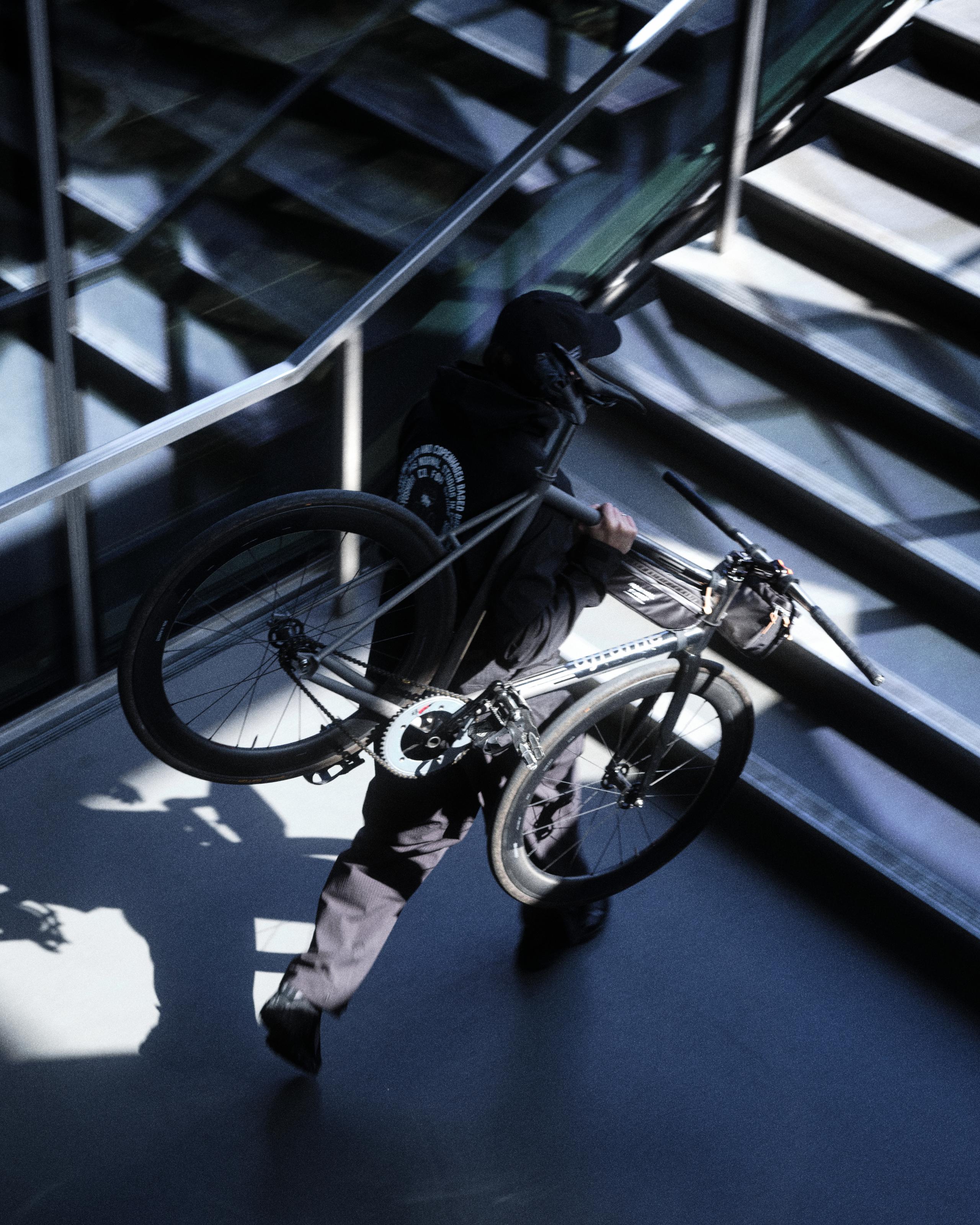 Model wearing black cycling kit carrying bike up stairs in dramatic shadows