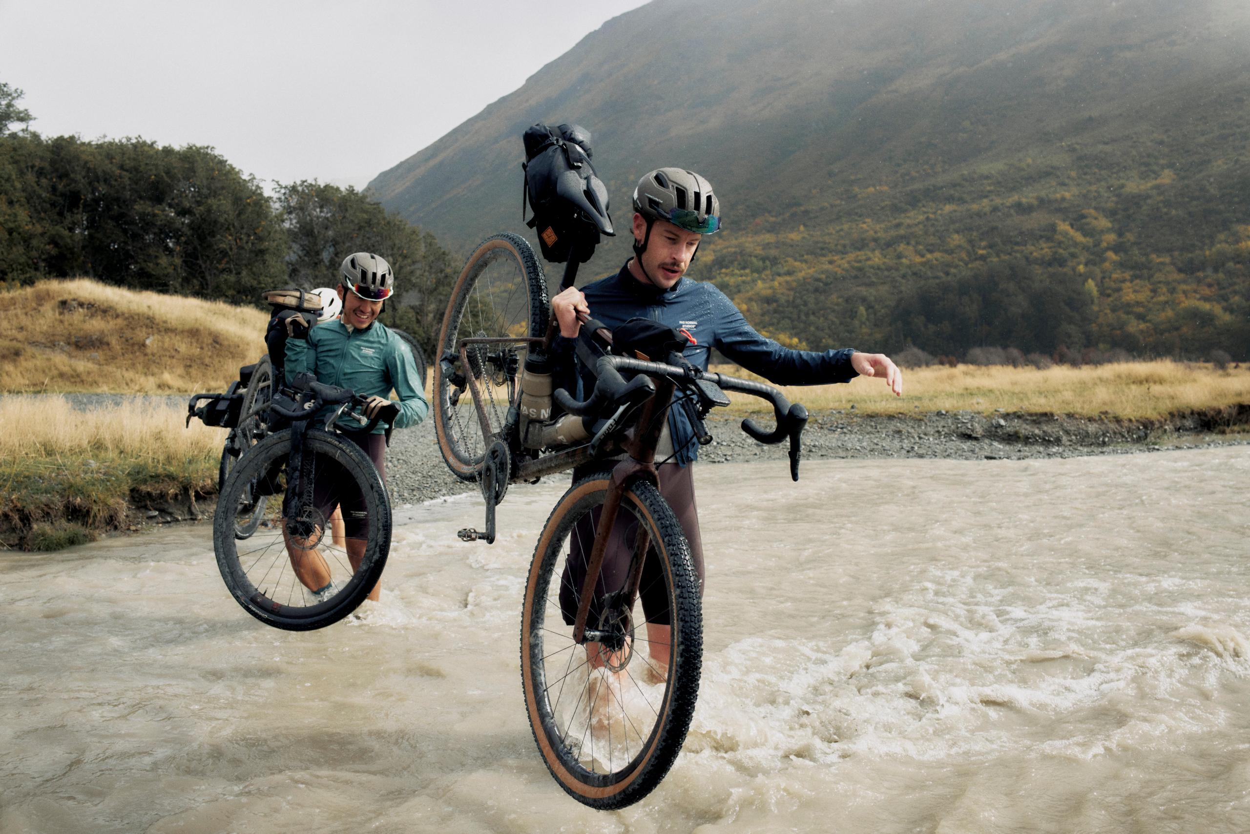 Two cyclists with gravel bikes crossing a stream in mountainous terrain, wearing cycling apparel and helmets