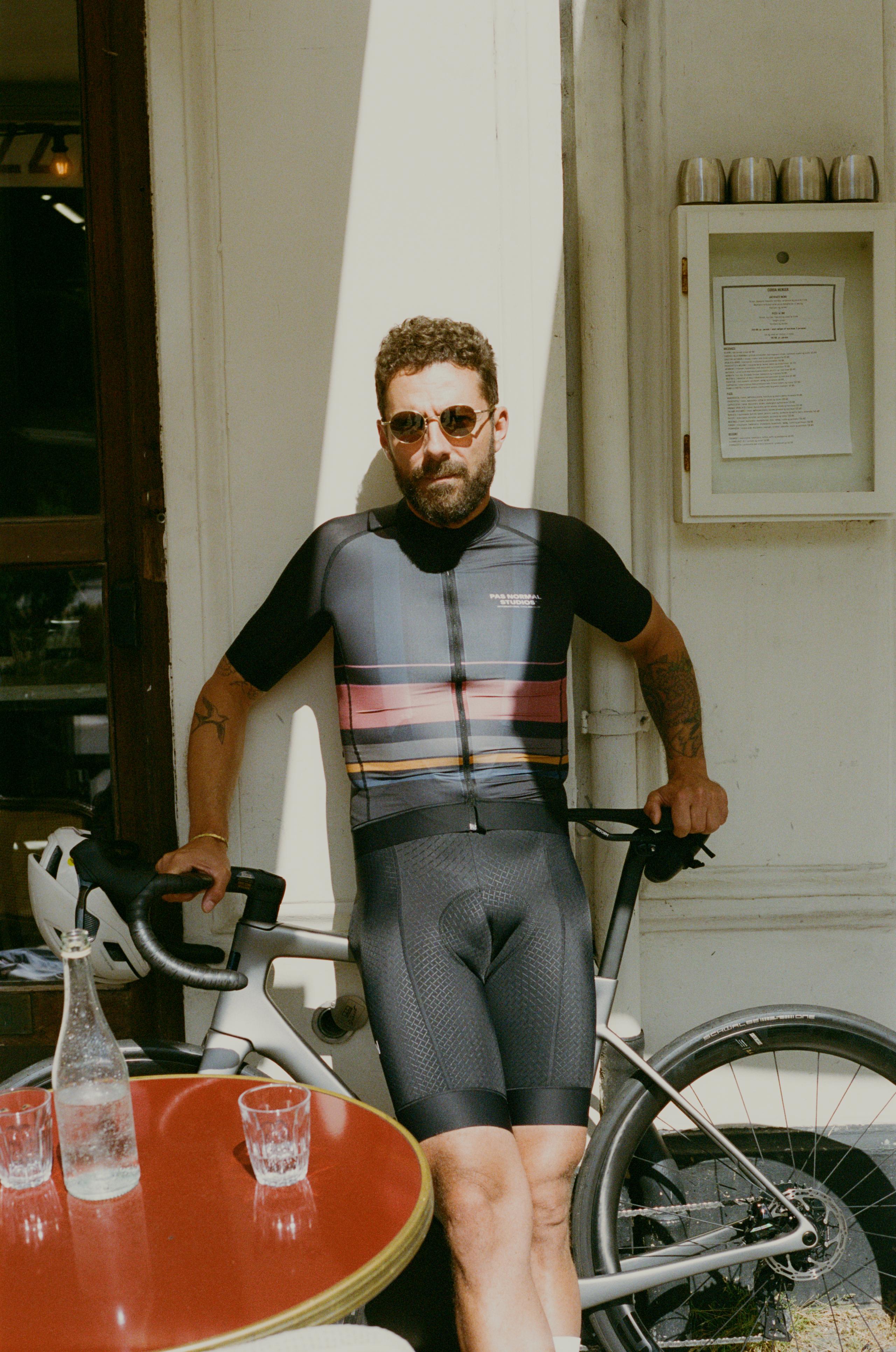 Model wearing Men's Mechanism skinsuit in grey with multicolored horizontal stripes, standing with bike at cafe