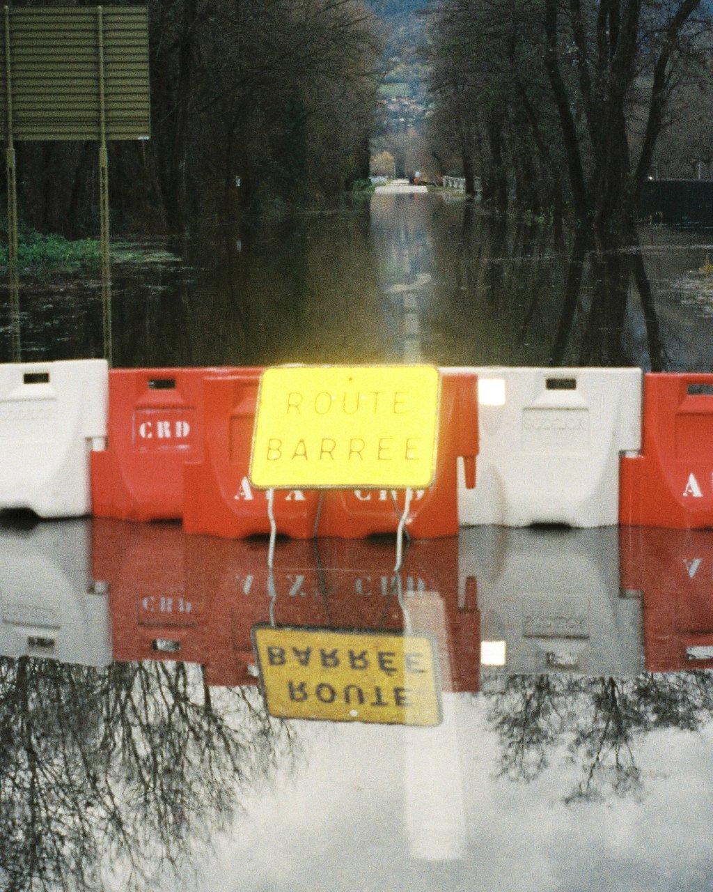 Flooded road blocked by orange and white barriers with yellow road closure sign reflected in water