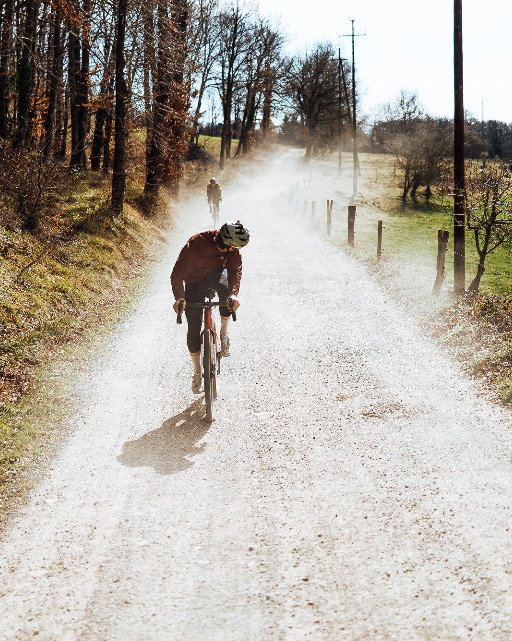 Cyclist in brown jersey riding gravel road with dust clouds on rural countryside route