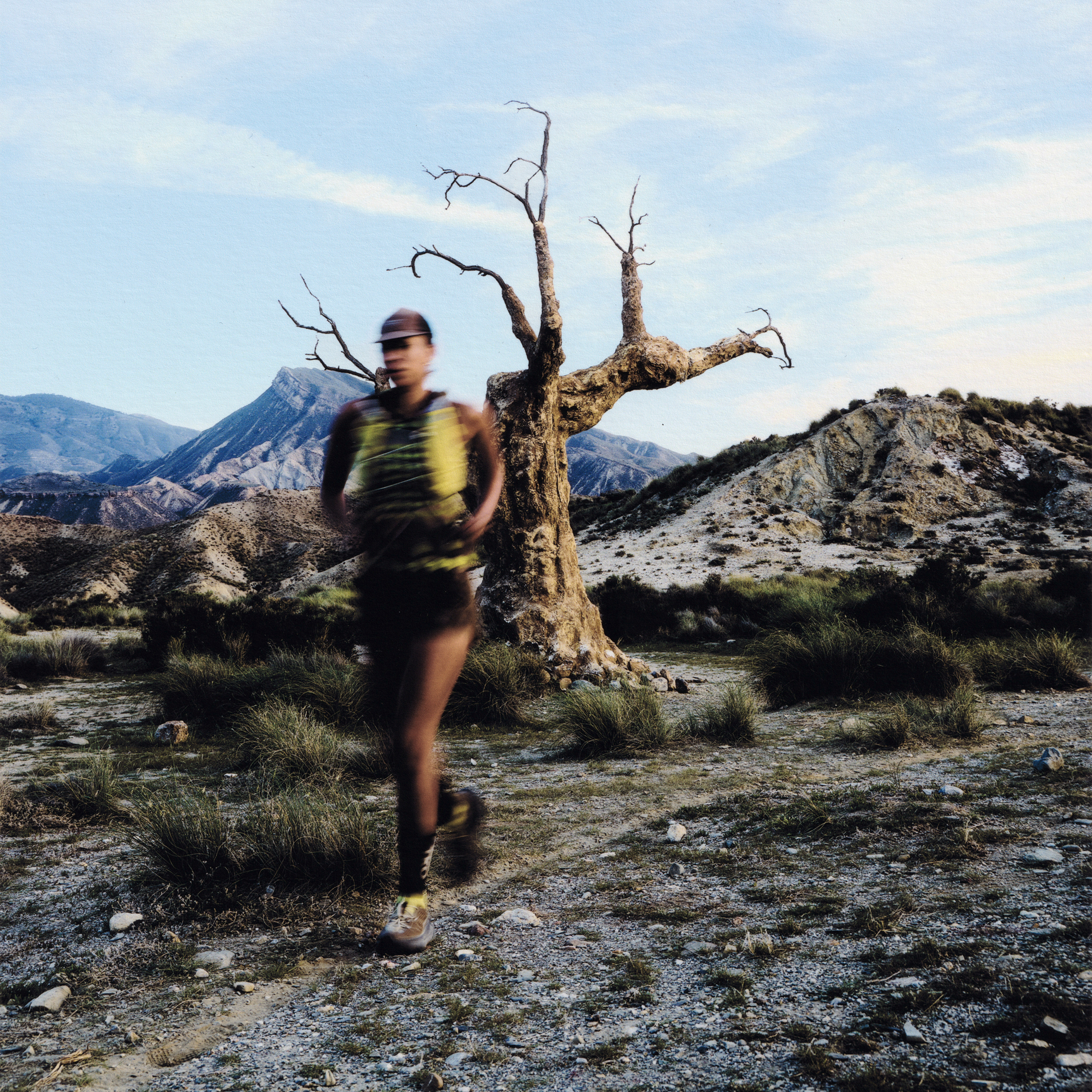 Model wearing green and black running gear in motion through desert landscape with dead tree and mountains