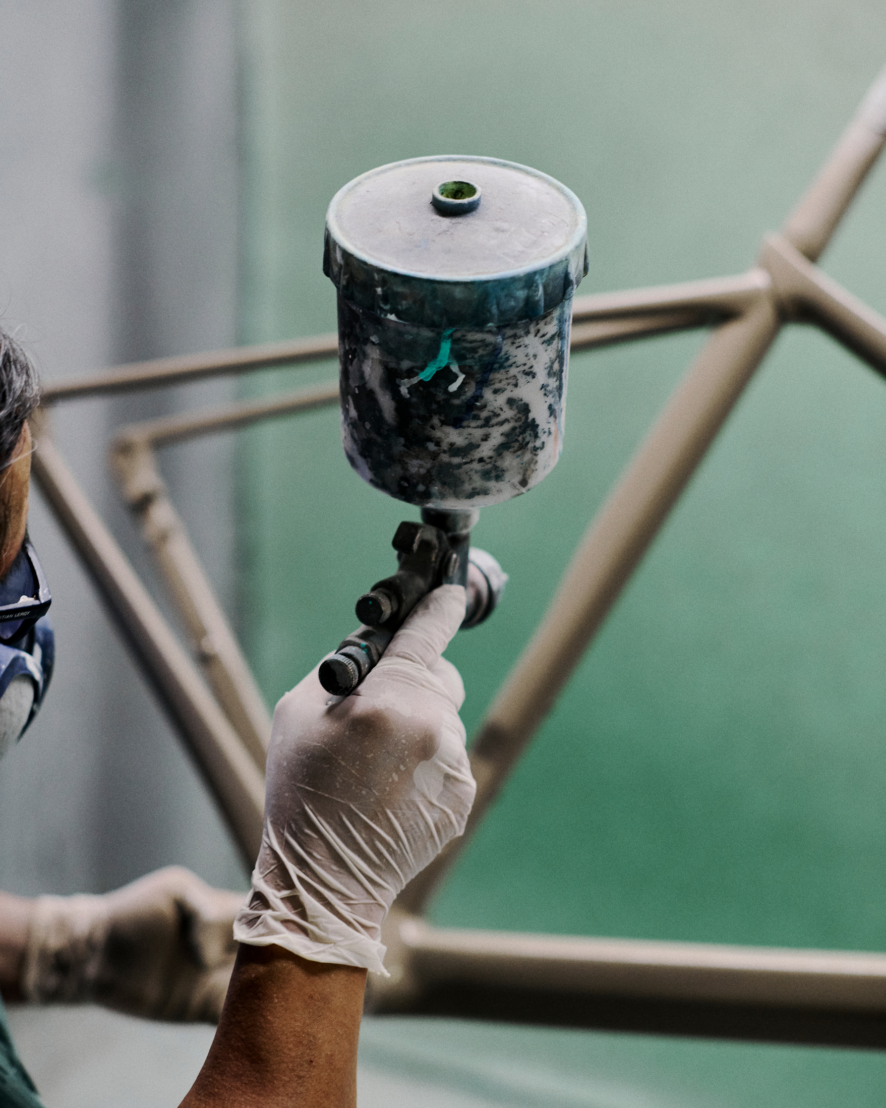 Hand holding paint sprayer with dark green and white splattered finish in workshop setting