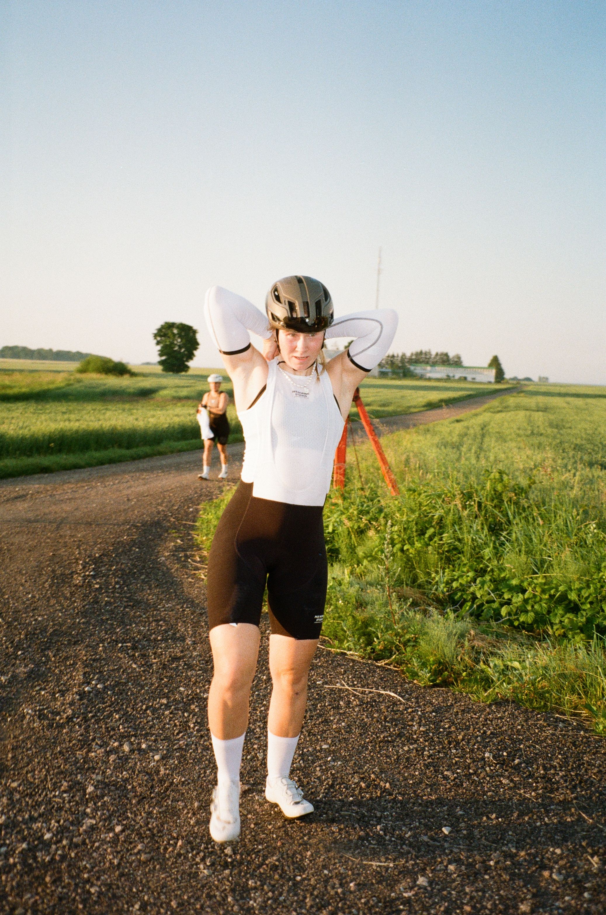 Model wearing white sleeveless cycling jersey and black shorts with time trial helmet on country road