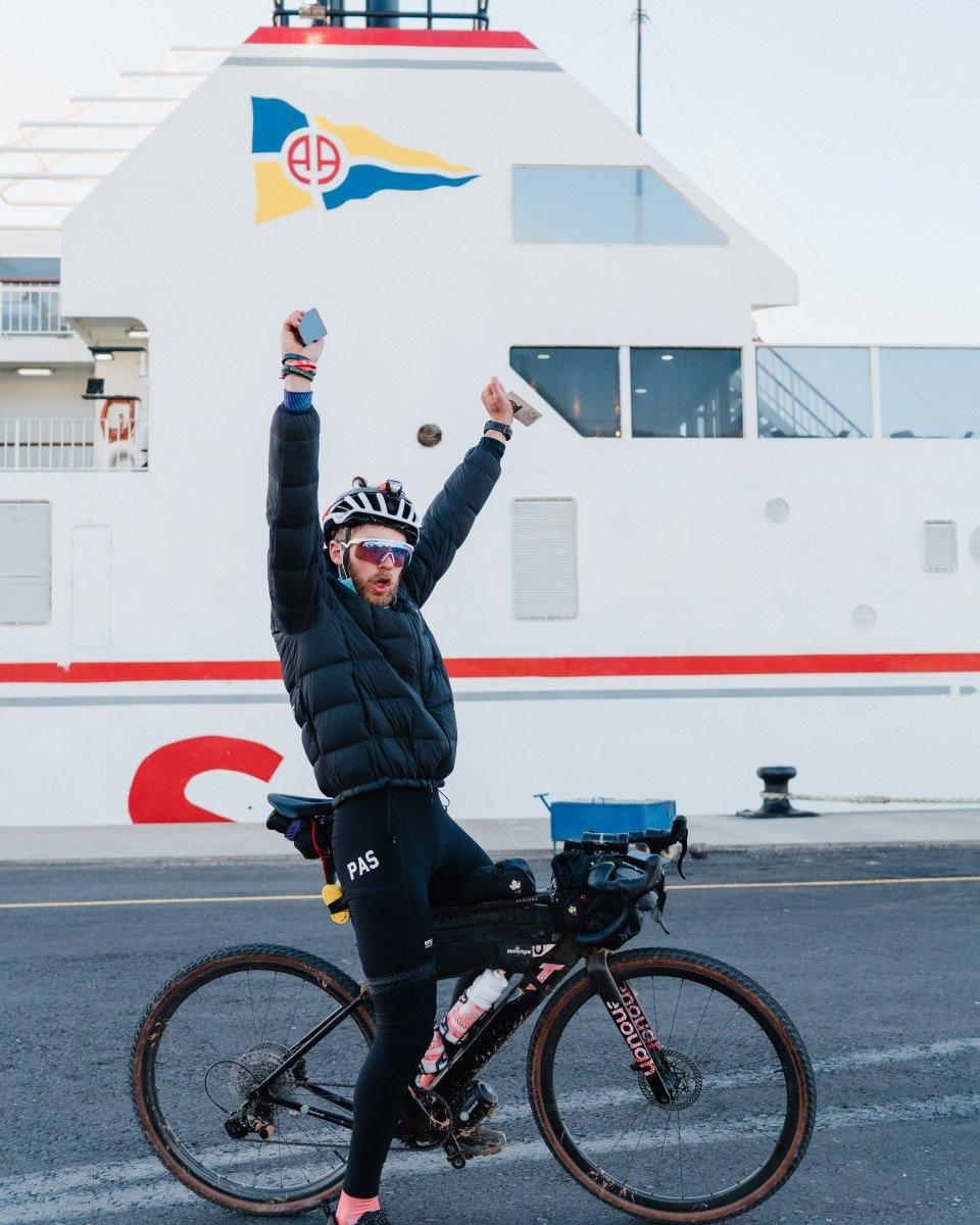 Model wearing black insulated jacket and PAS bib tights celebrating with arms raised on bike near ferry
