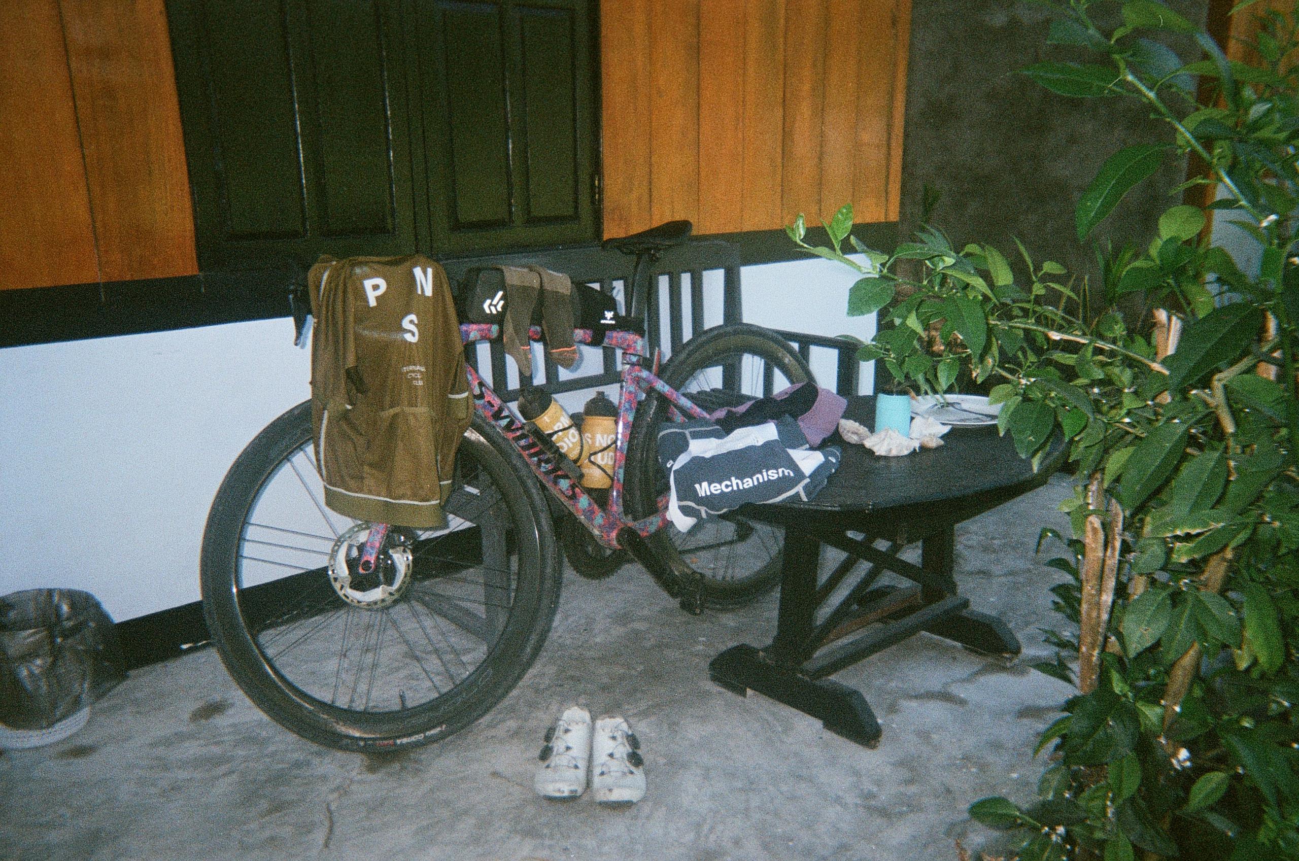 Cycling gear and accessories arranged on wooden bench with bicycle, including brown jersey with logo and patterned items