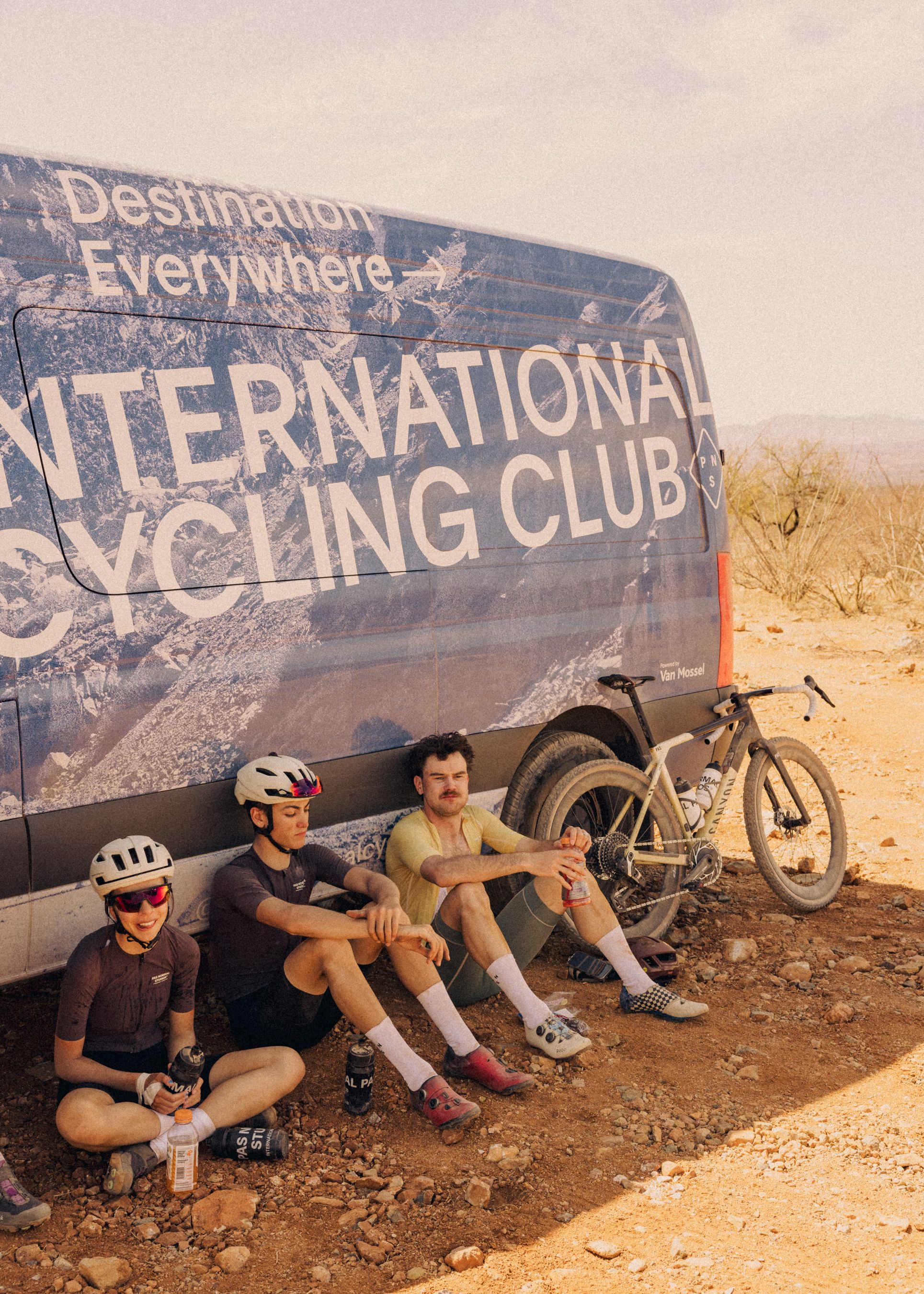 Three cyclists resting in desert setting beside International Cycling Club van with mountain bikes