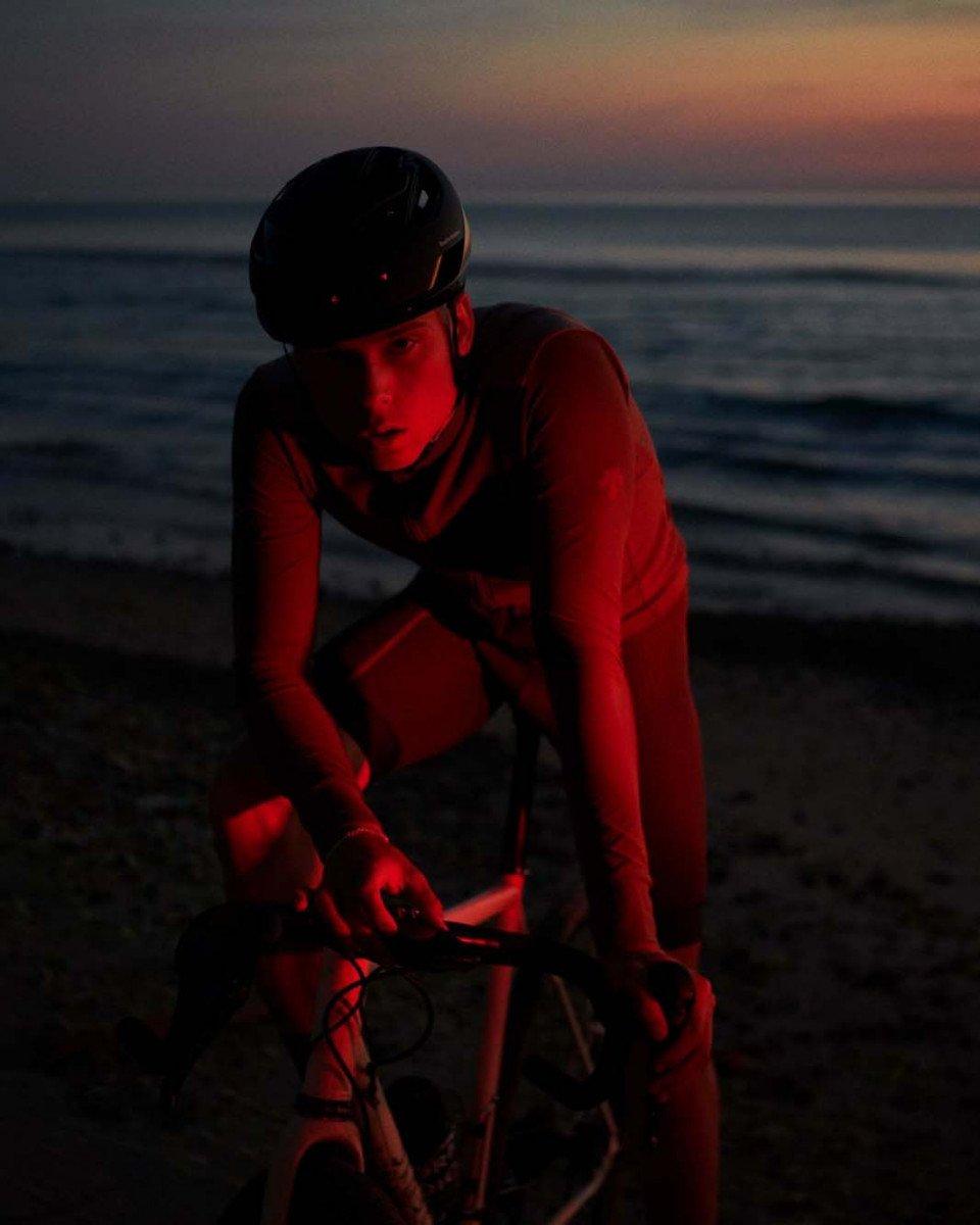 Model wearing black helmet and cycling gear with red lighting by ocean at sunset