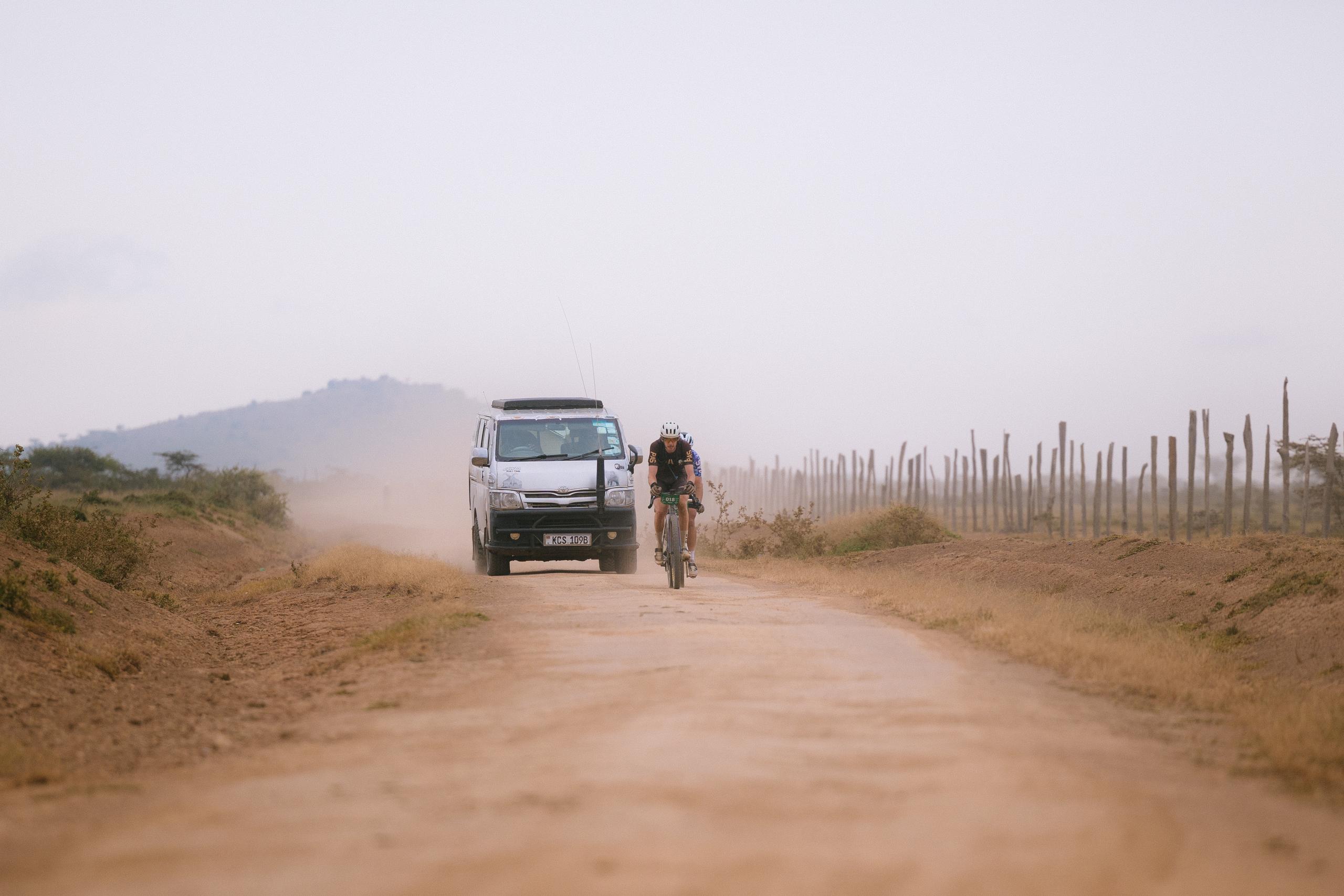 Cyclist riding on dusty gravel road with vehicle support in rural landscape