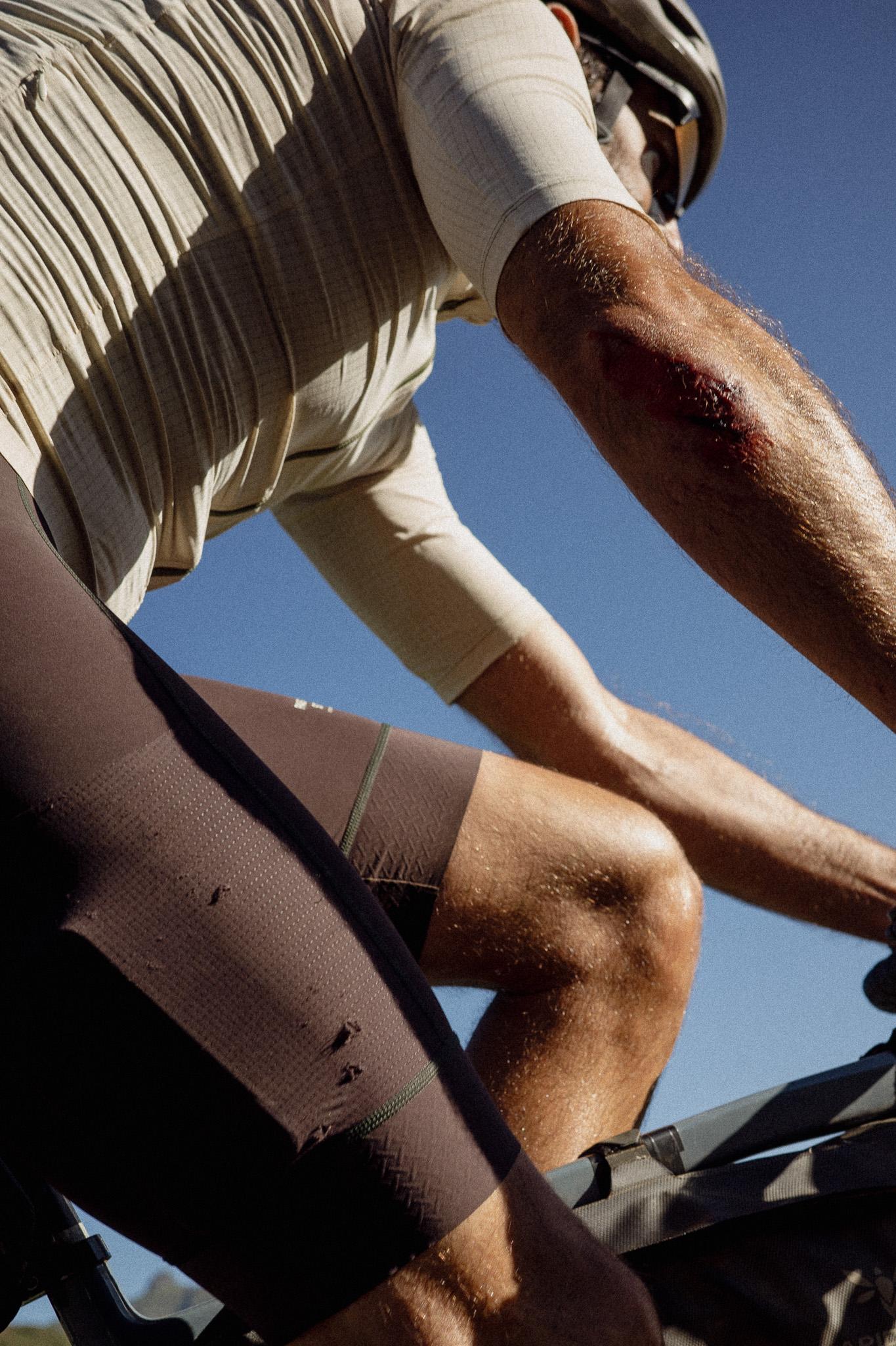Model wearing beige jersey and brown shorts with green trim, cycling under blue sky