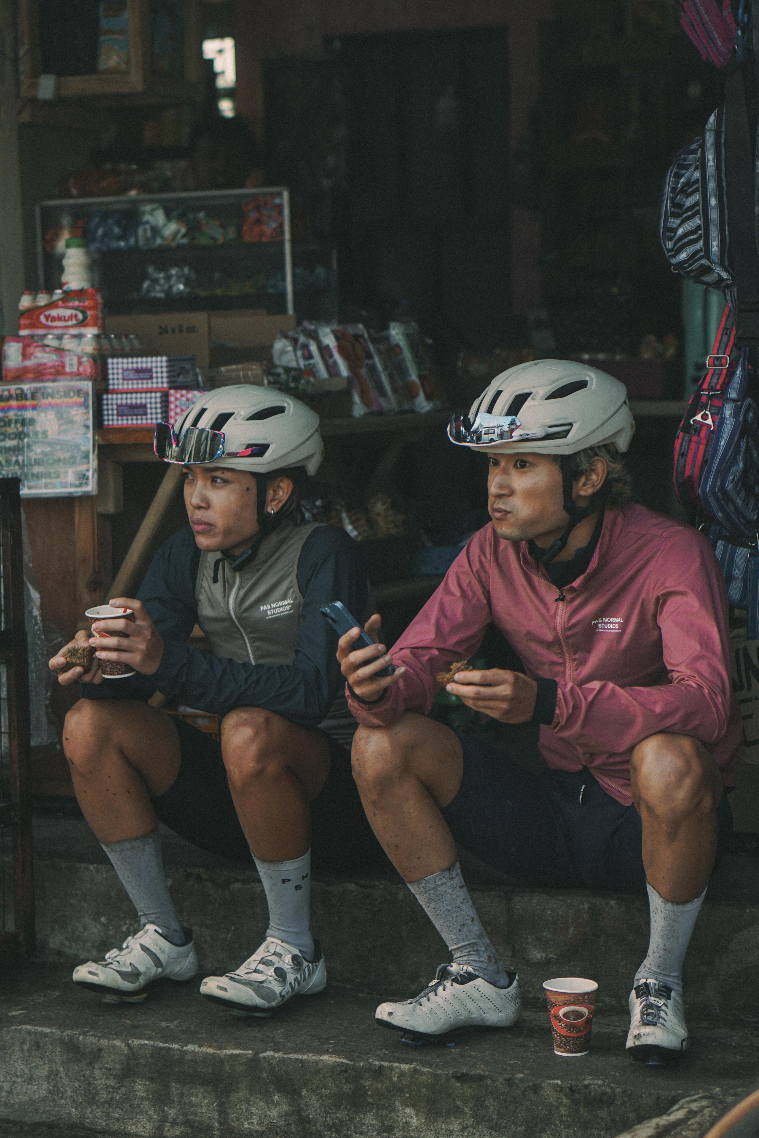 Two cyclists in helmets wearing long-sleeve jerseys, one olive green and one burgundy, resting at roadside shop
