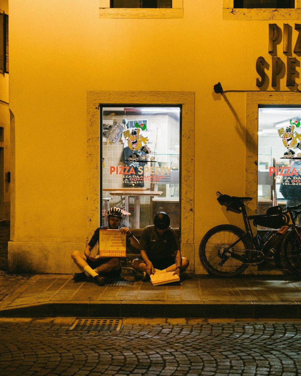 Two cyclists resting outside pizza shop at night with bicycle parked nearby