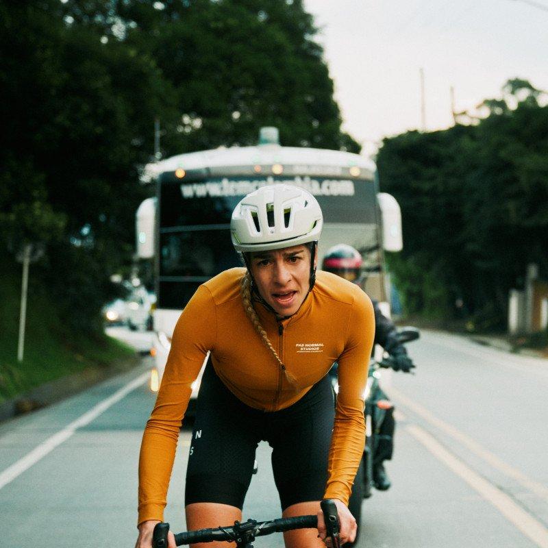 Model wearing orange long-sleeve jersey and black shorts cycling on urban road with bus in background