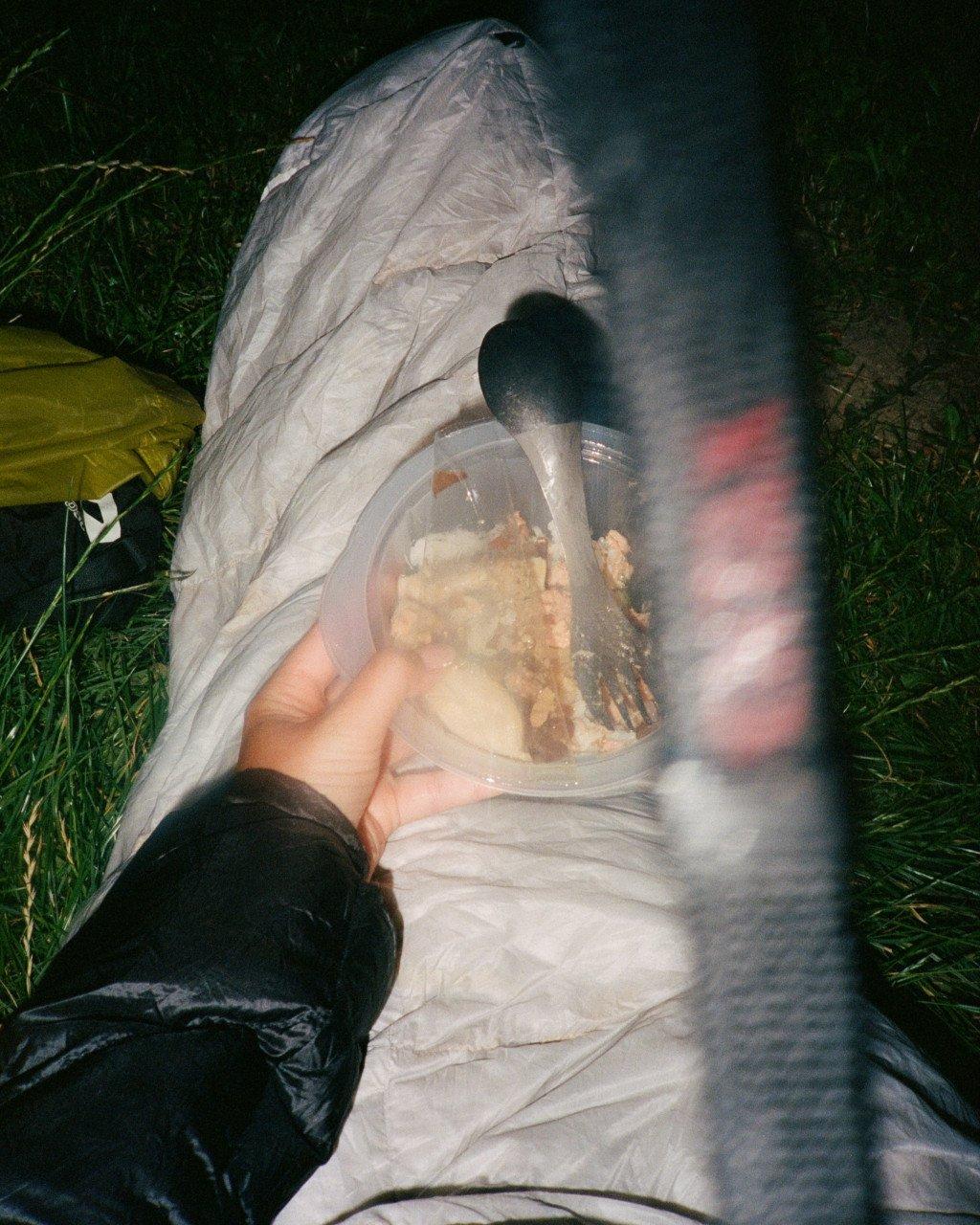 Person in sleeping bag inside tent holding glass of food while camping