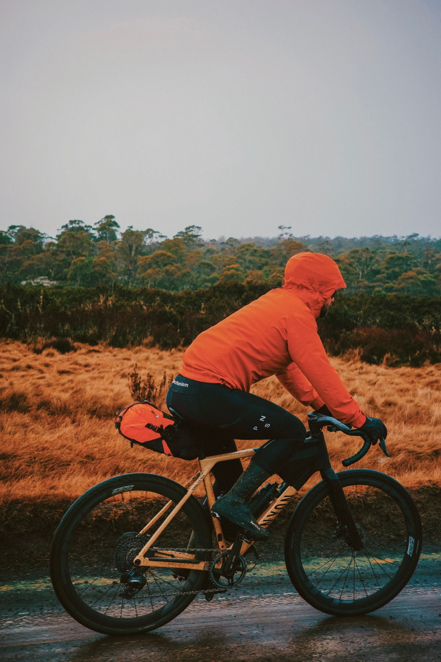 Model wearing orange hoodie and black cycling shorts on time trial bike in natural landscape