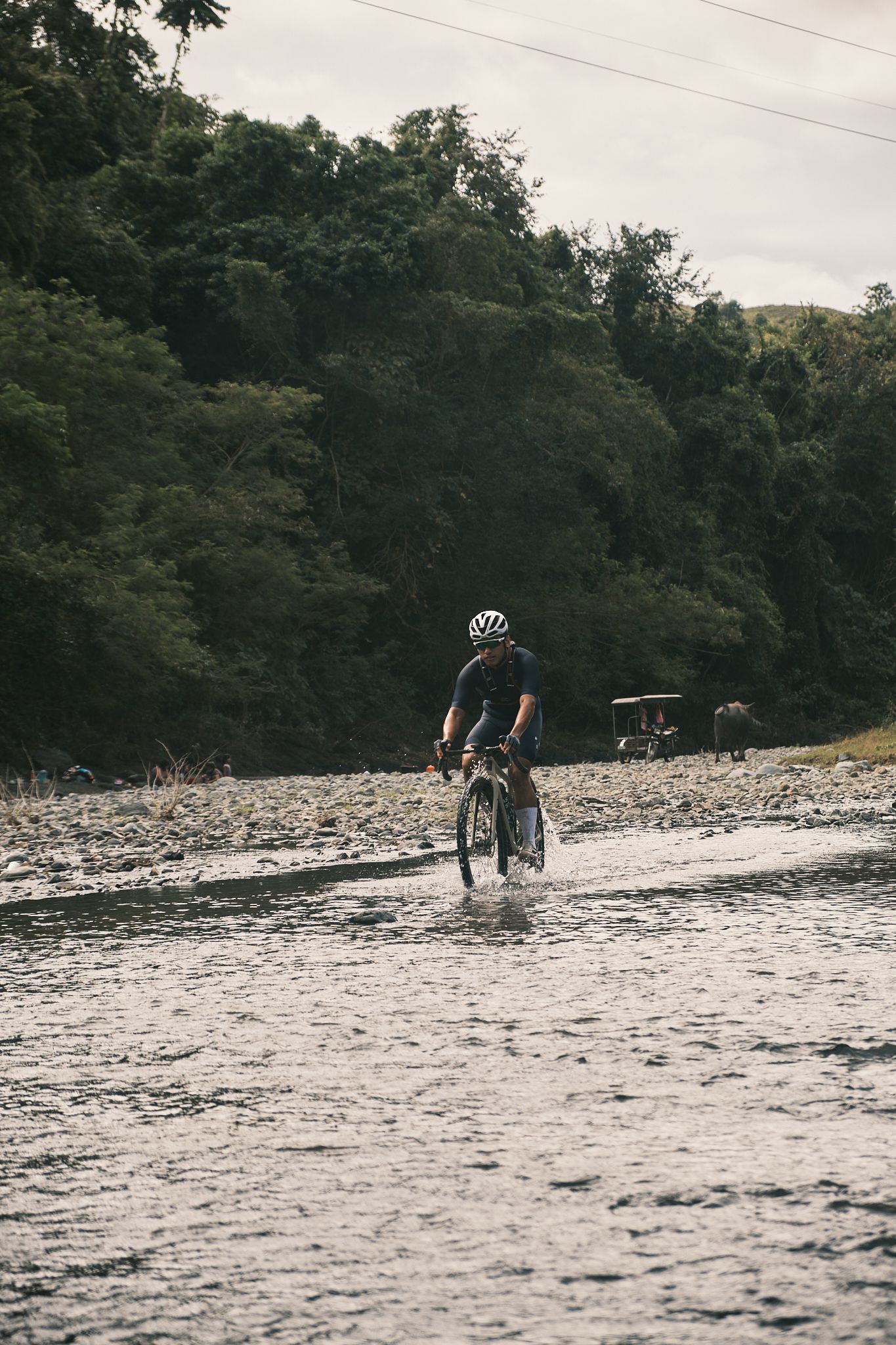Cyclist in dark clothing riding through shallow water on rocky riverbed with forested hills in background