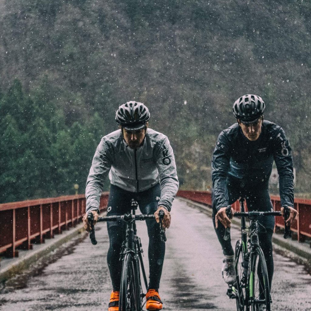 Two cyclists riding through rain on a bridge, wearing dark cycling jackets and helmets