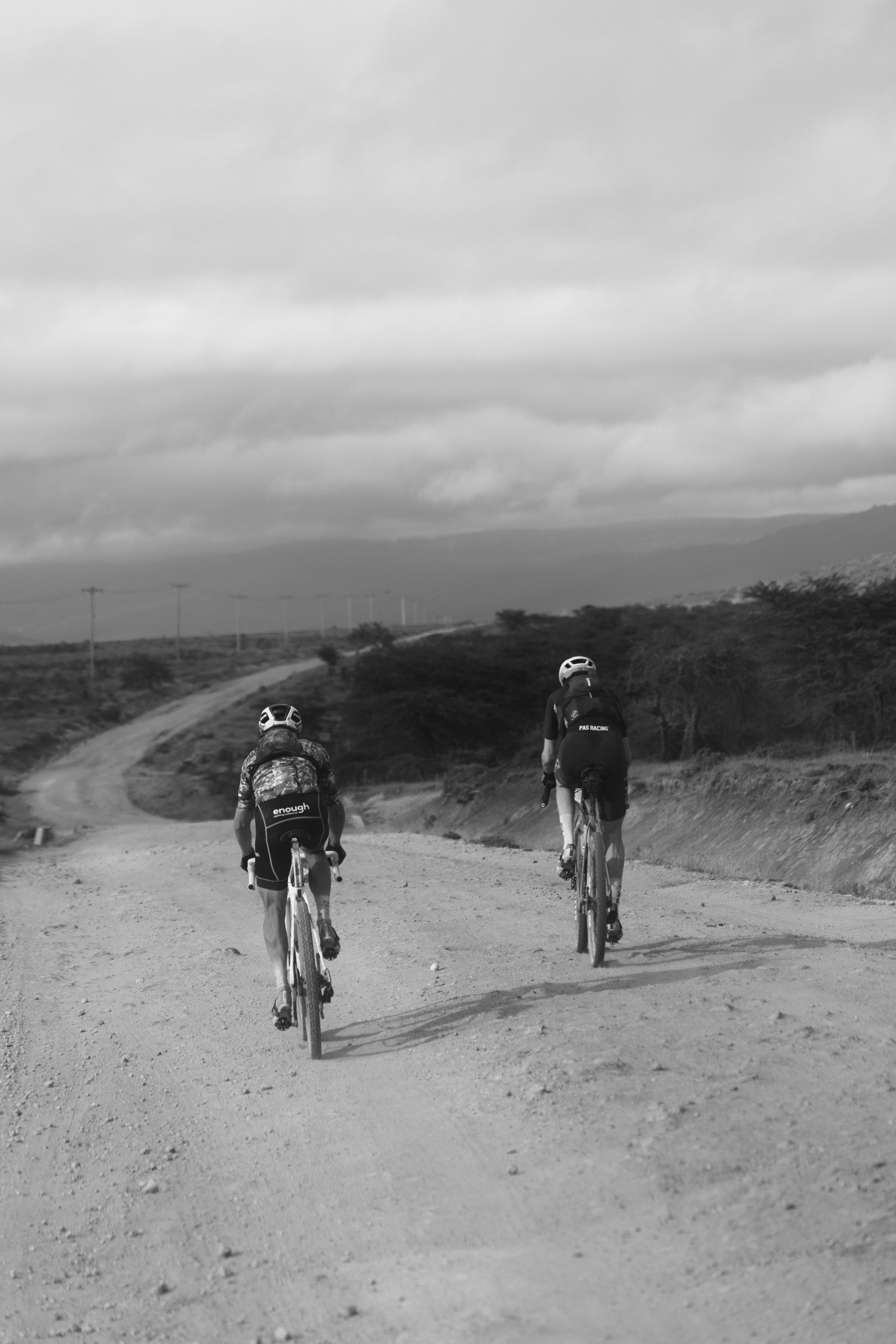 Two cyclists riding gravel bikes on dirt road through desert landscape in black and white