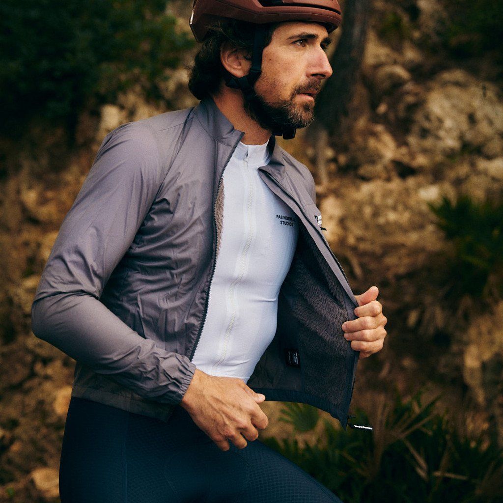 Model wearing grey zip-up cycling jacket with dark interior, white tee underneath, helmet in outdoor setting