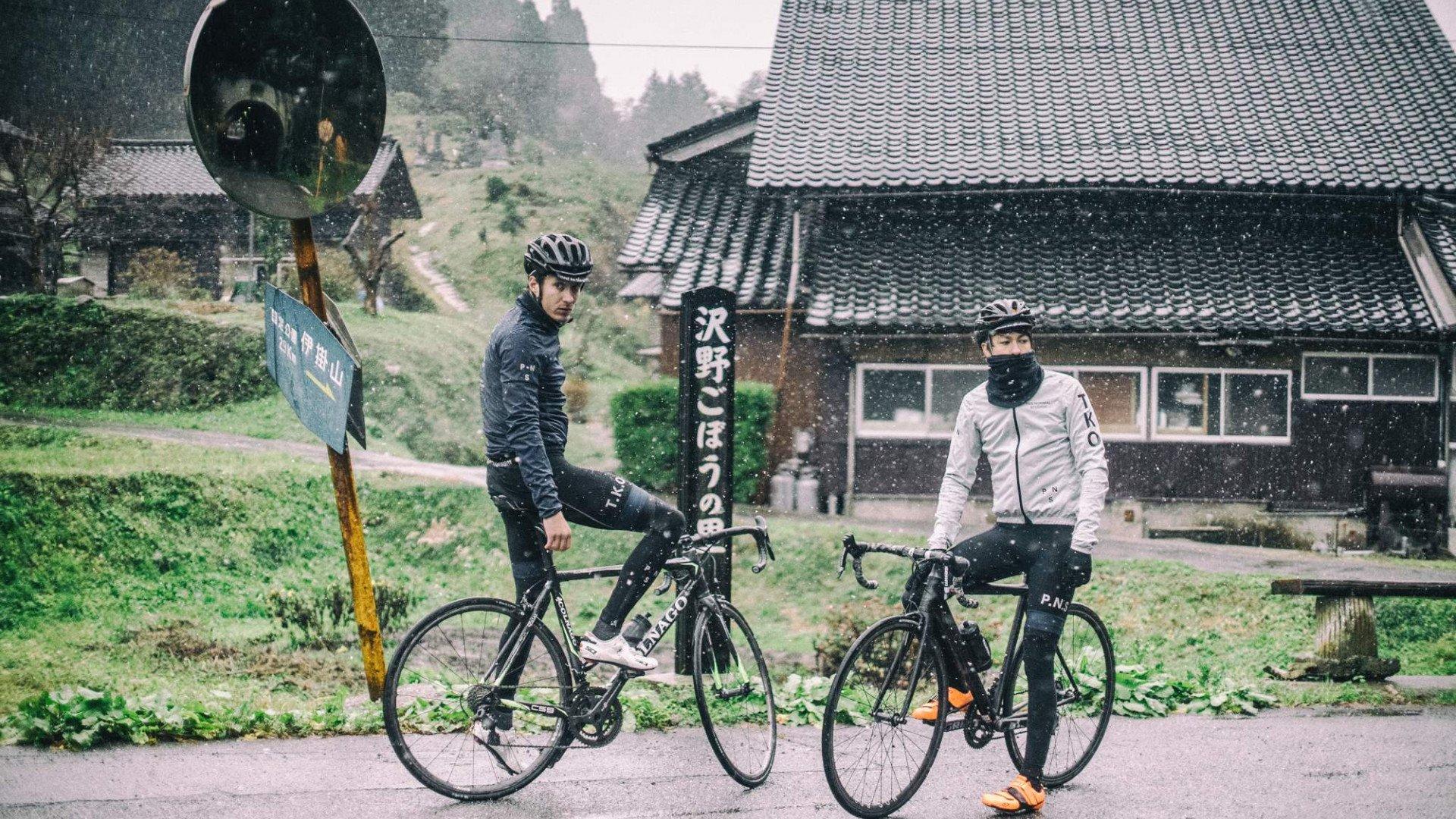 Two cyclists with bikes in snowy Japanese village, one in dark jacket, one in light jacket with neck warmer