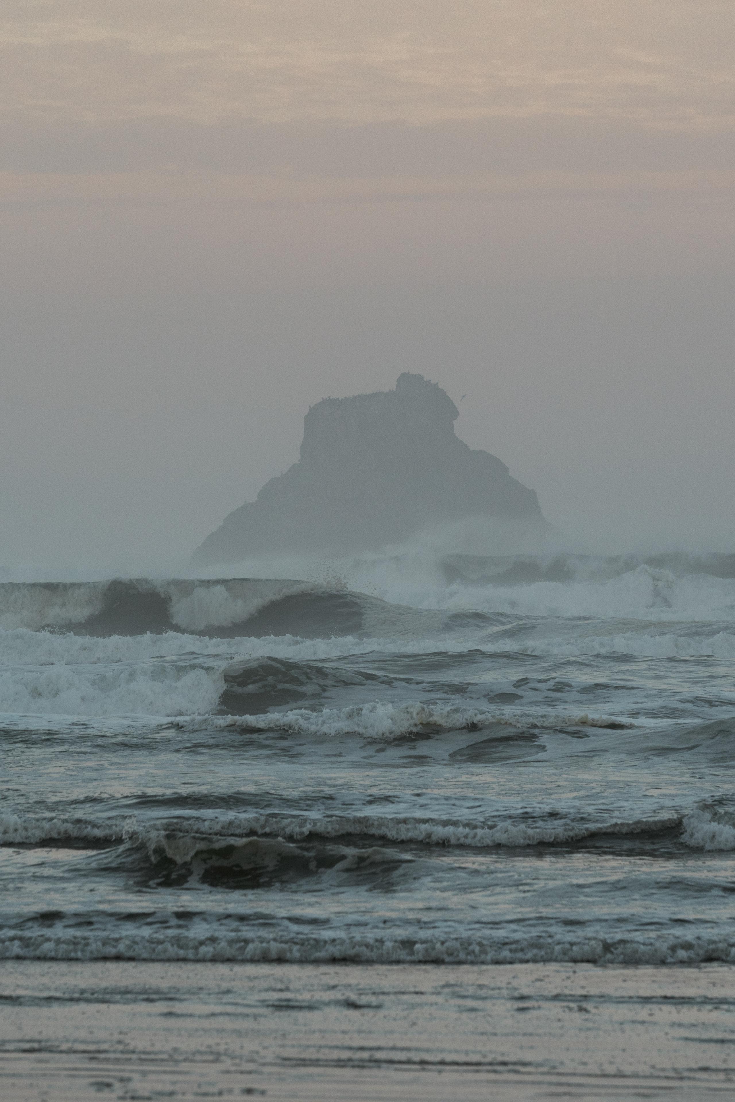 Misty coastal landscape with waves breaking on beach and rocky outcrop in fog