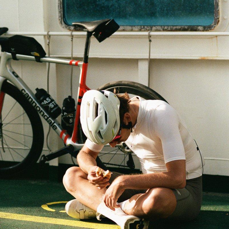 Model wearing white jersey and helmet sitting cross-legged near bicycle in garage setting