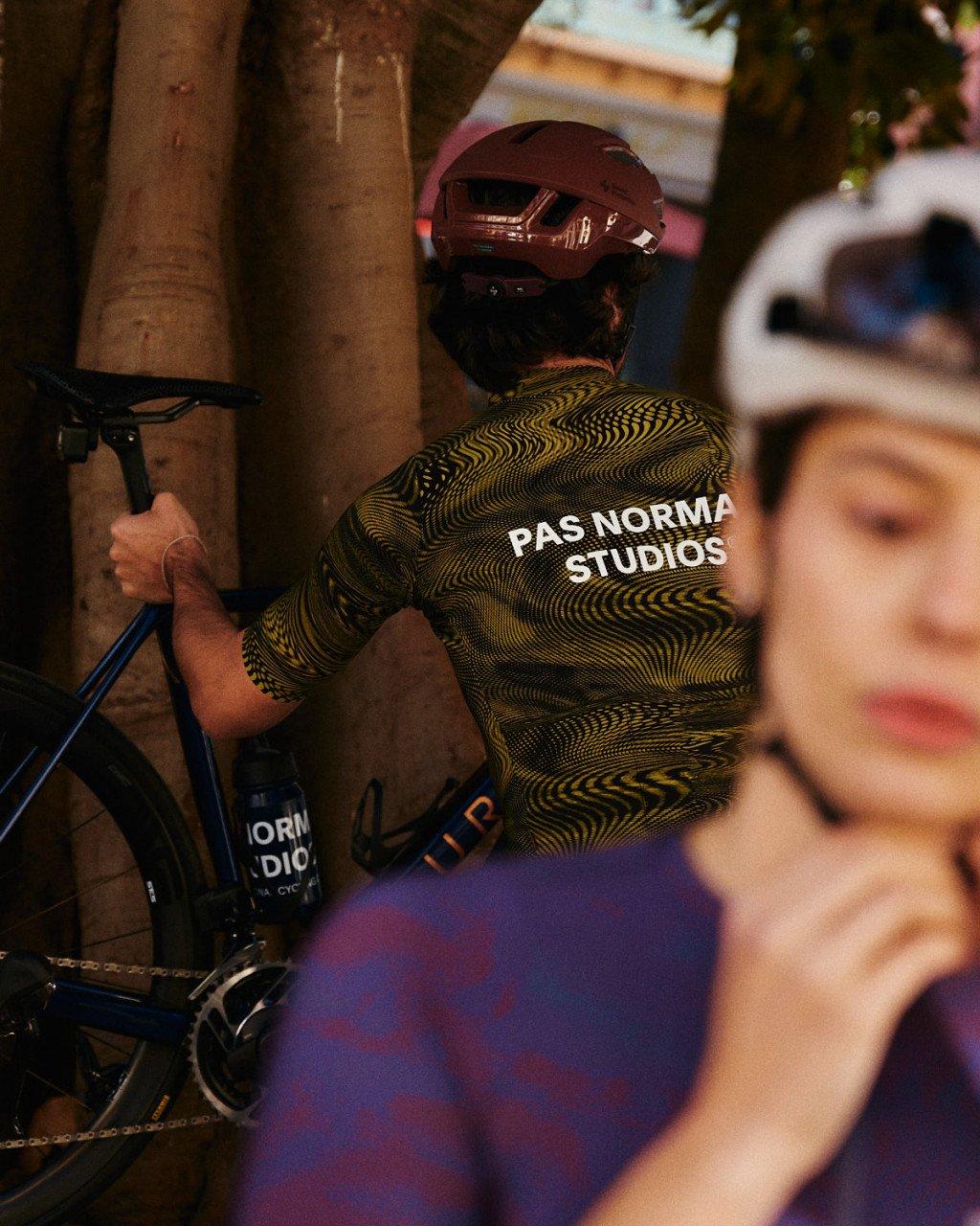Model wearing yellow and black geometric patterned cycling jersey with back branding, burgundy helmet, urban setting