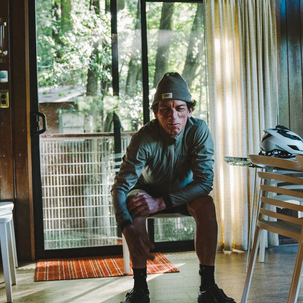 Model wearing grey beanie and green cycling jacket sitting indoors with cycling helmet on table