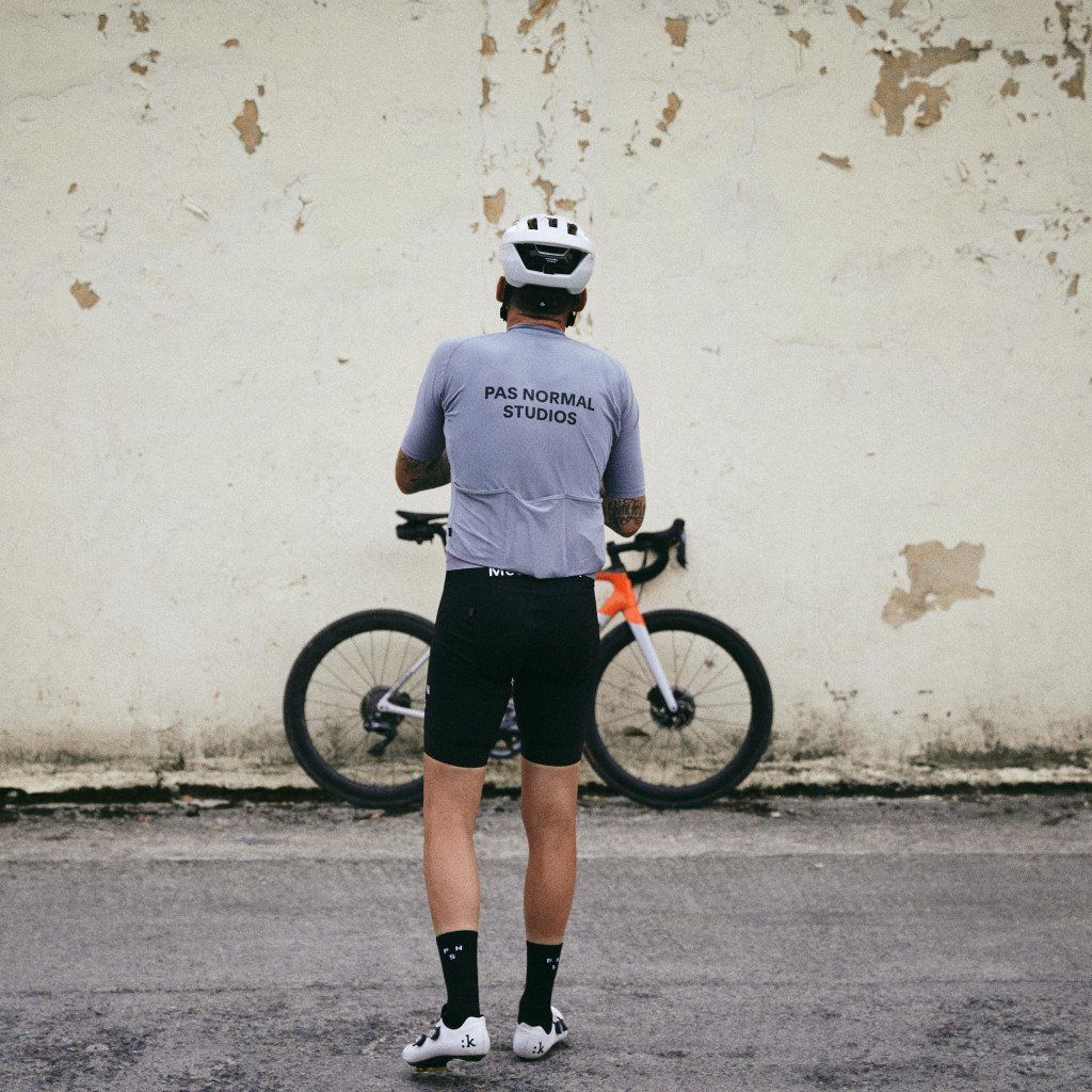 Model wearing light purple jersey with back branding and black cycling shorts, standing with bike against weathered wall