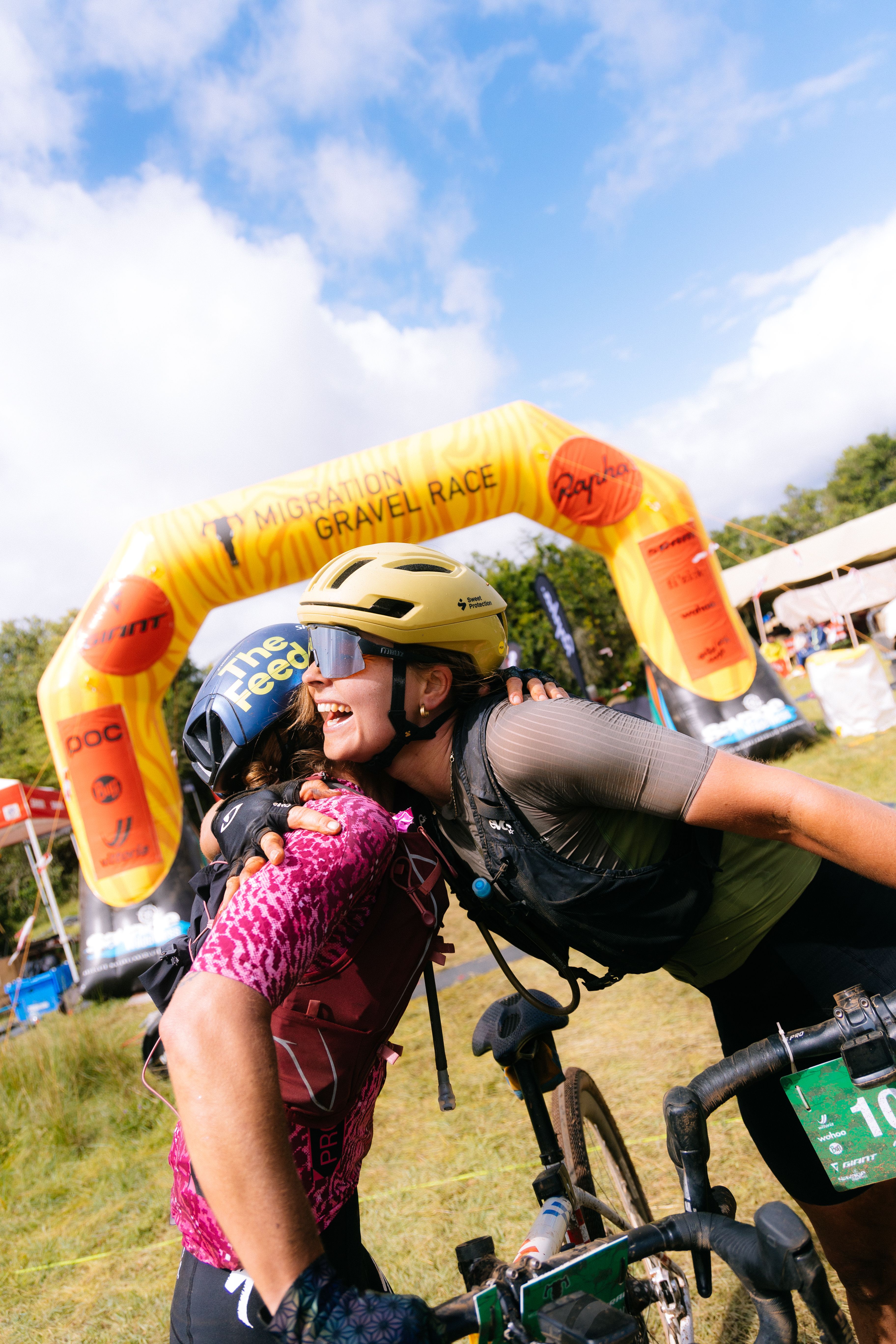 Female cyclists embracing at gravel race finish line with inflatable arch and bikes