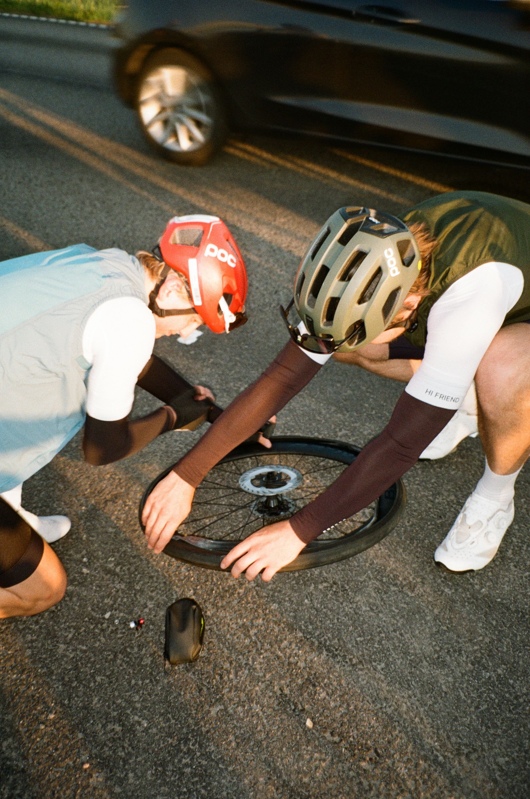Two cyclists fixing a bike wheel on road, wearing helmets and cycling apparel