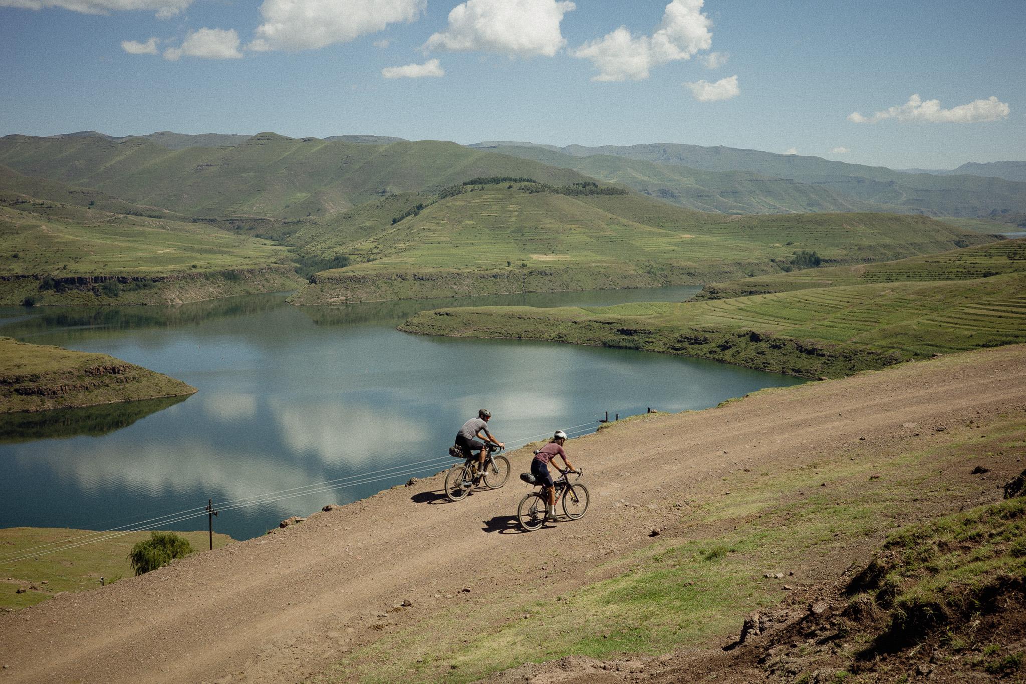 Two cyclists riding gravel bikes on dirt road beside mountain lake in Lesotho highlands