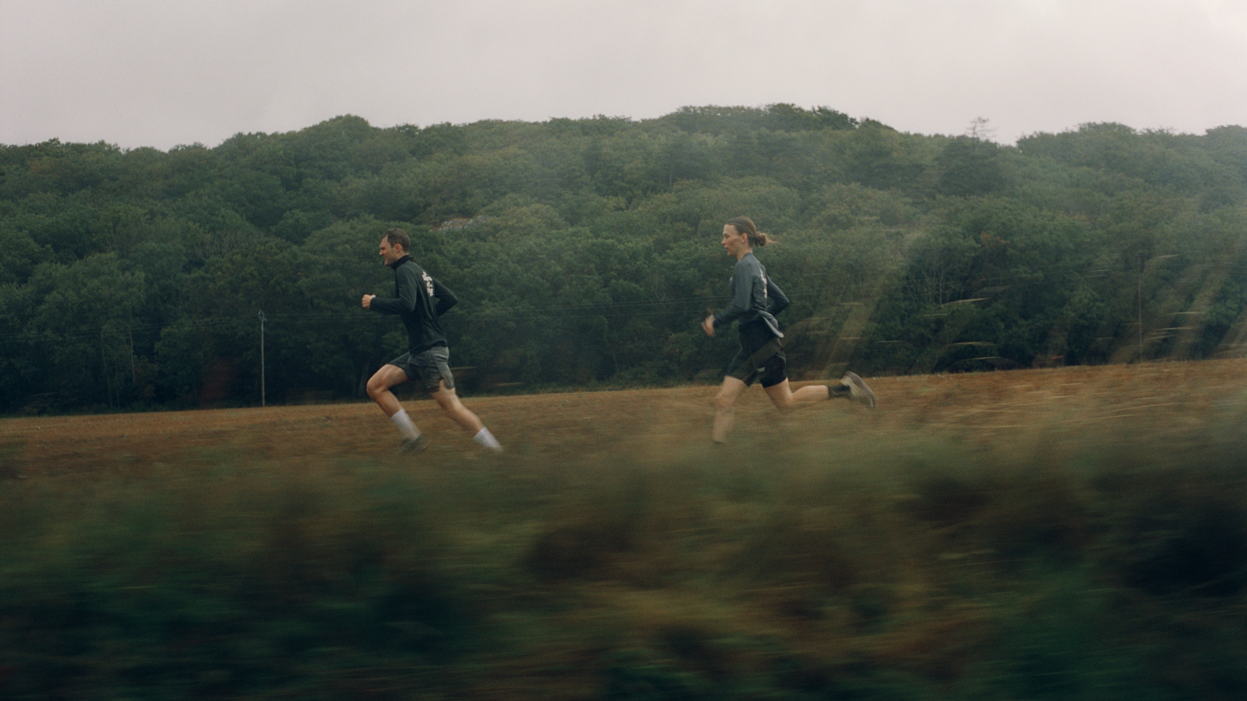 Two runners in dark athletic wear running across open field with forested hills in background