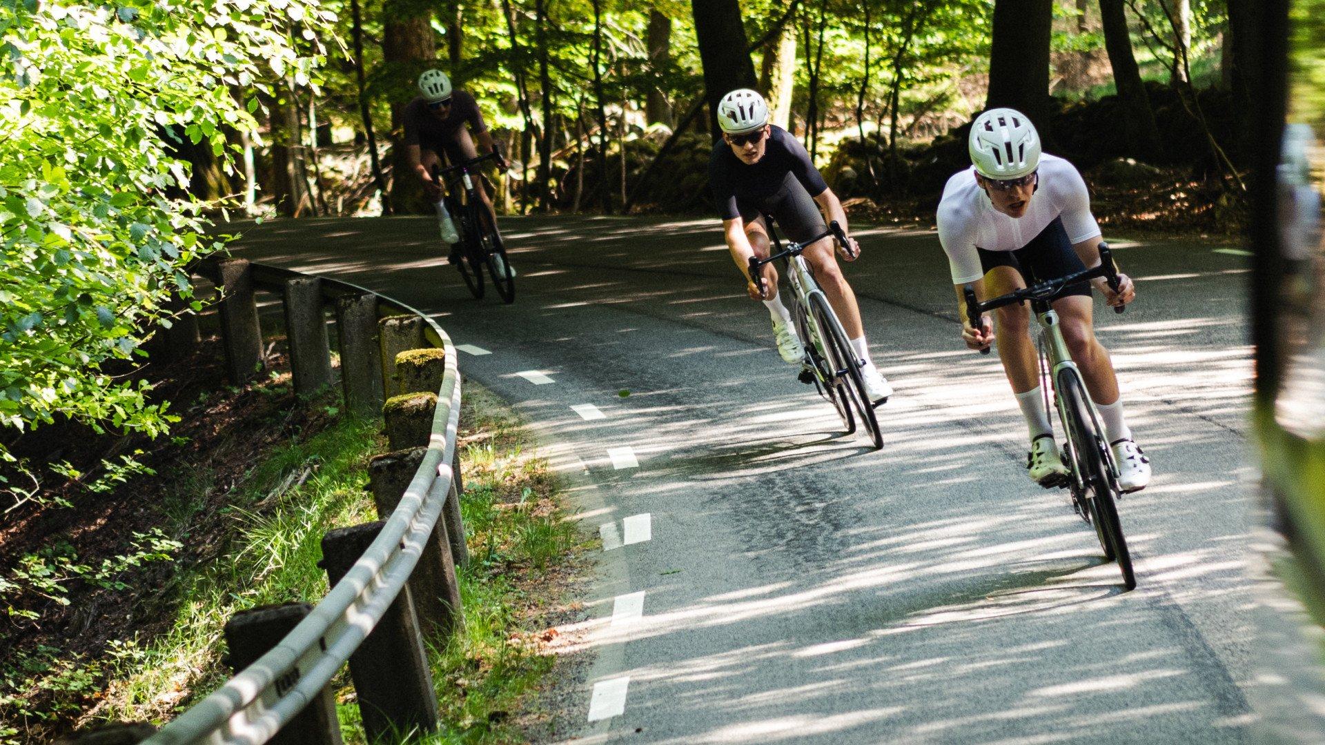 Two cyclists in aerodynamic position racing on forest road, one in black skinsuit, one in white skinsuit