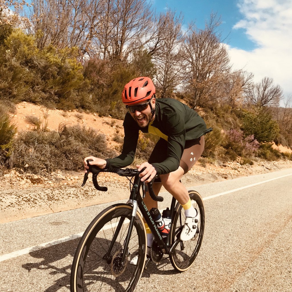 Model wearing olive green long sleeve cycling jersey with red helmet on time trial bike on desert road