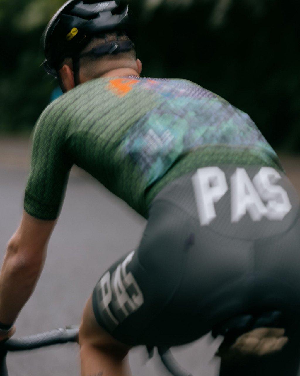 Model wearing green cycling jersey with multicolored pattern and collection lettering, shown from behind in cycling position
