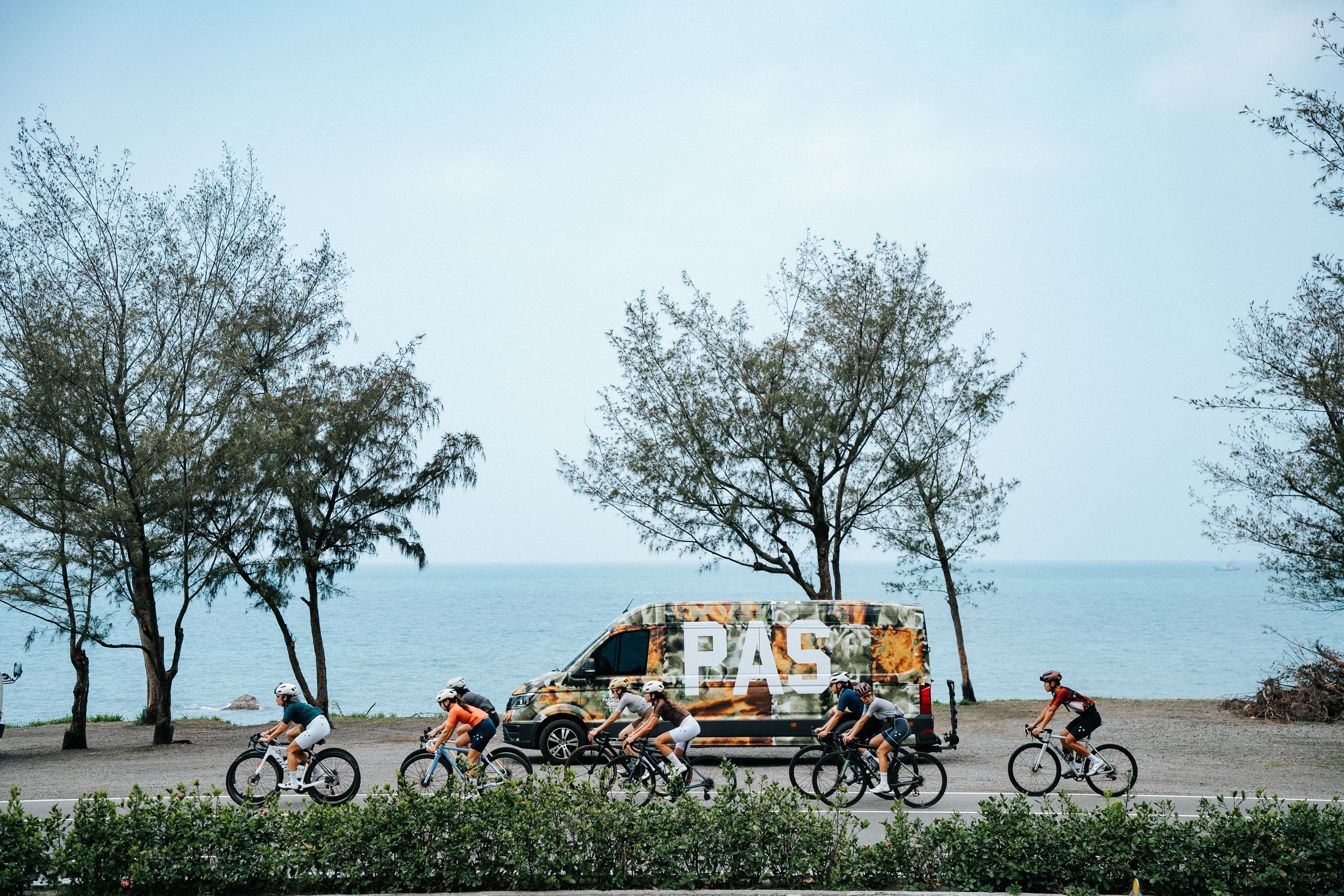 Cyclists riding past PAS branded support vehicle on coastal road with ocean and trees in background