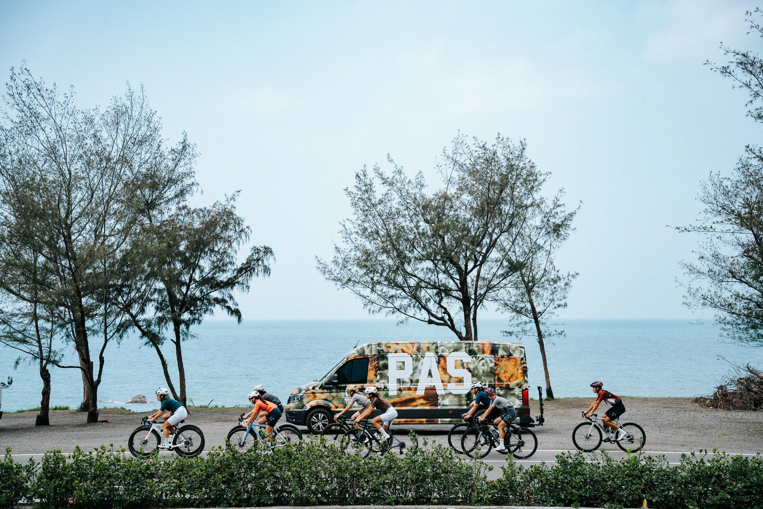 Cyclists riding past PAS branded support vehicle on coastal road with ocean and trees in background