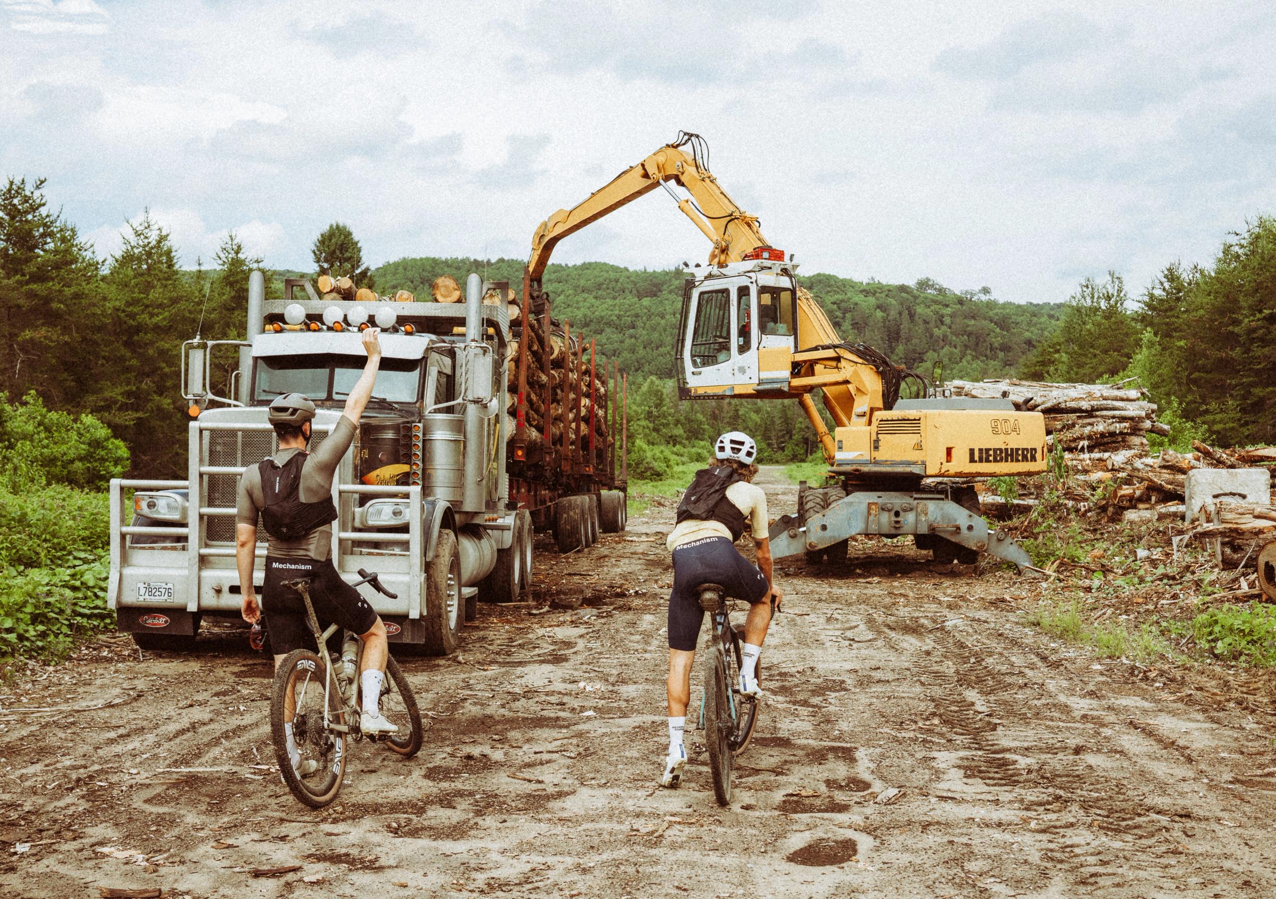 Two cyclists in dark jerseys and white helmets riding past construction equipment on dirt road