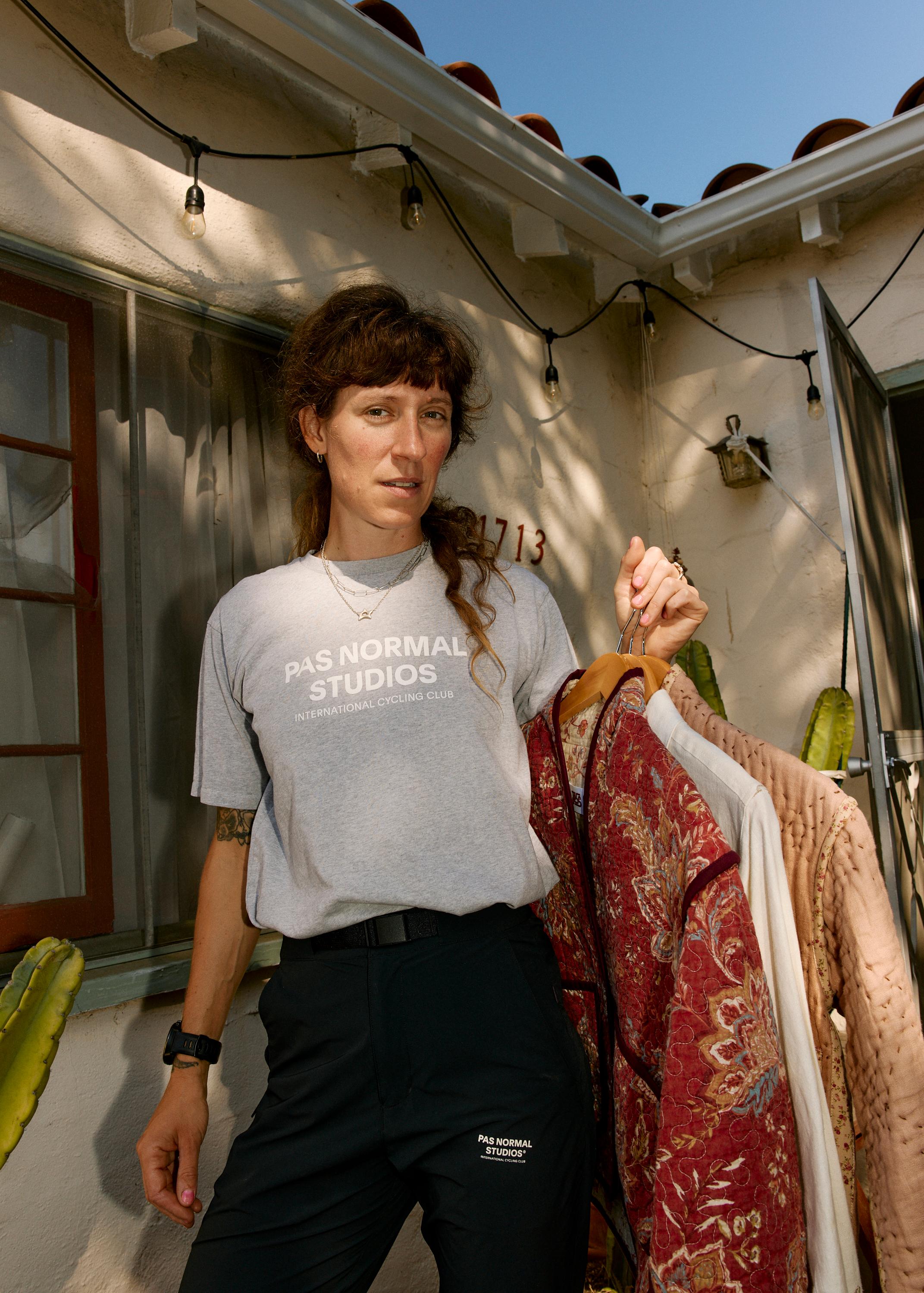 Model wearing grey t-shirt with chest lettering and dark pants, holding clothing on hangers in courtyard setting