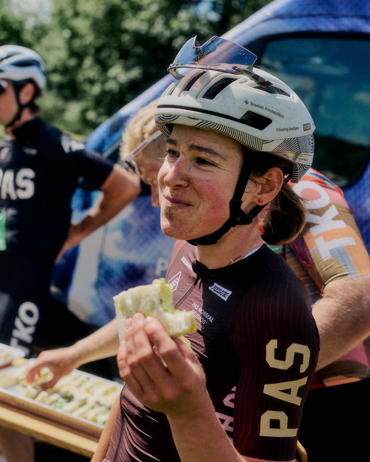 Female cyclist wearing maroon jersey with large text, white cycling helmet, eating at outdoor event
