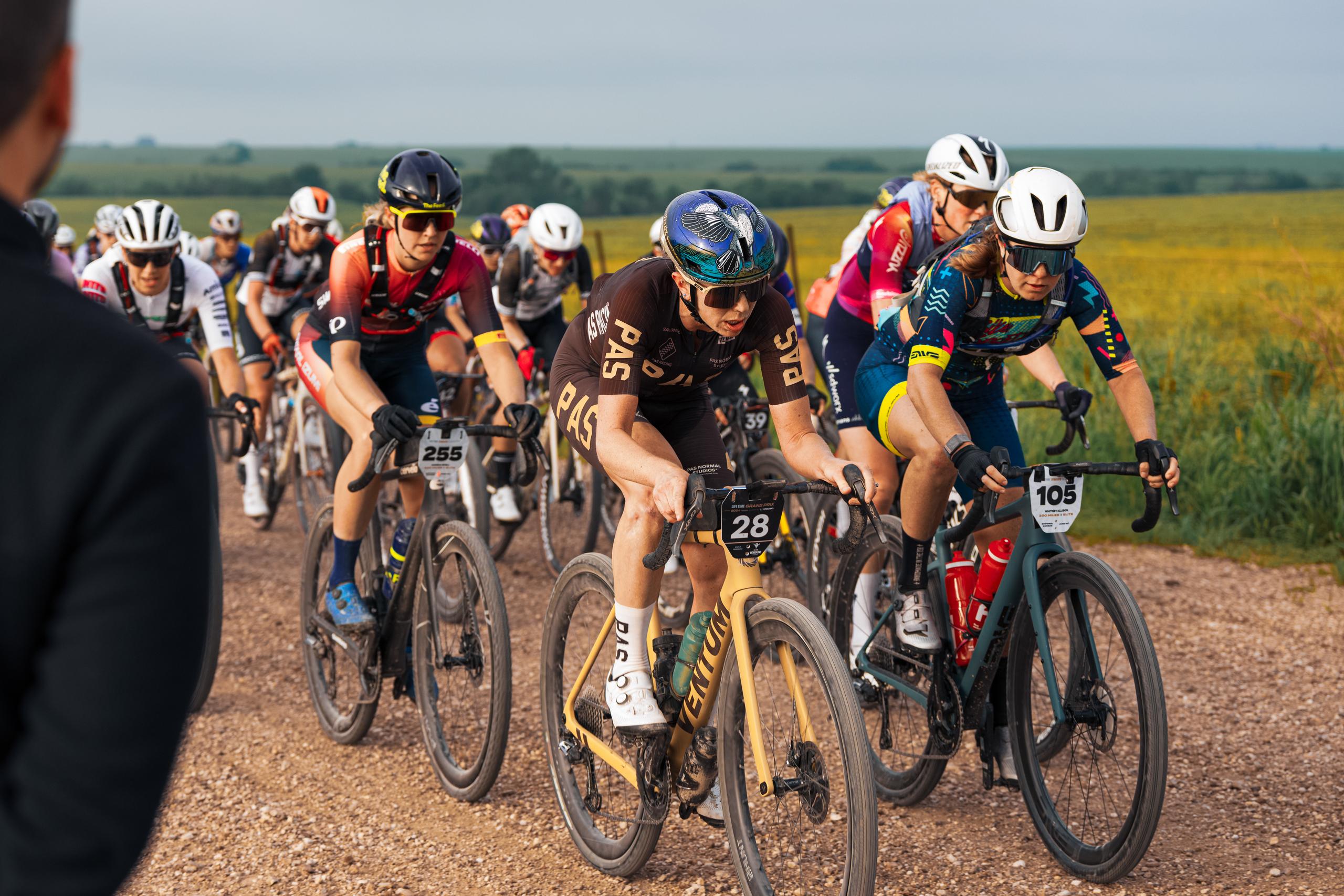 Cyclists racing on gravel road wearing various team jerseys and aero helmets