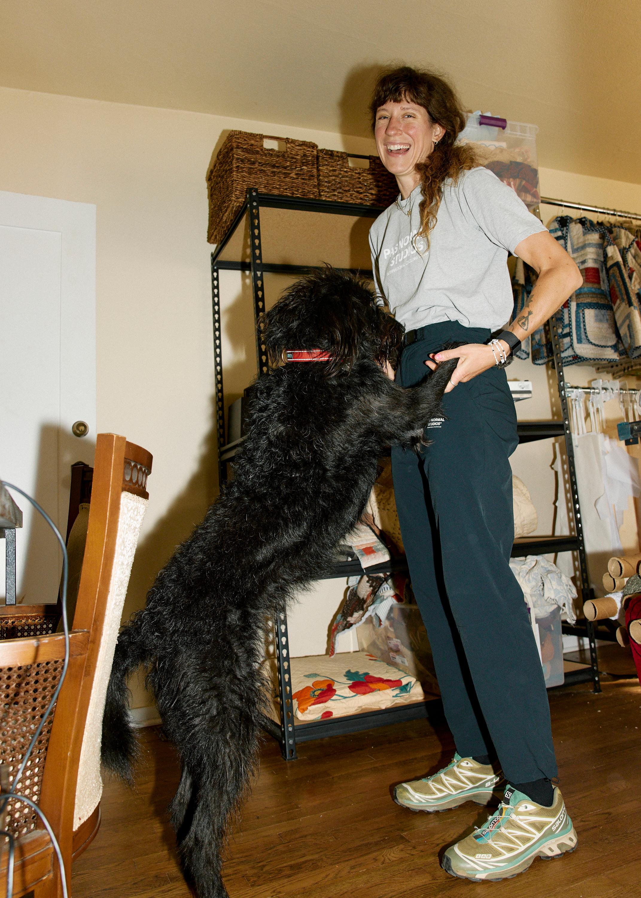 Model wearing grey t-shirt and navy trousers with large black dog in home office setting