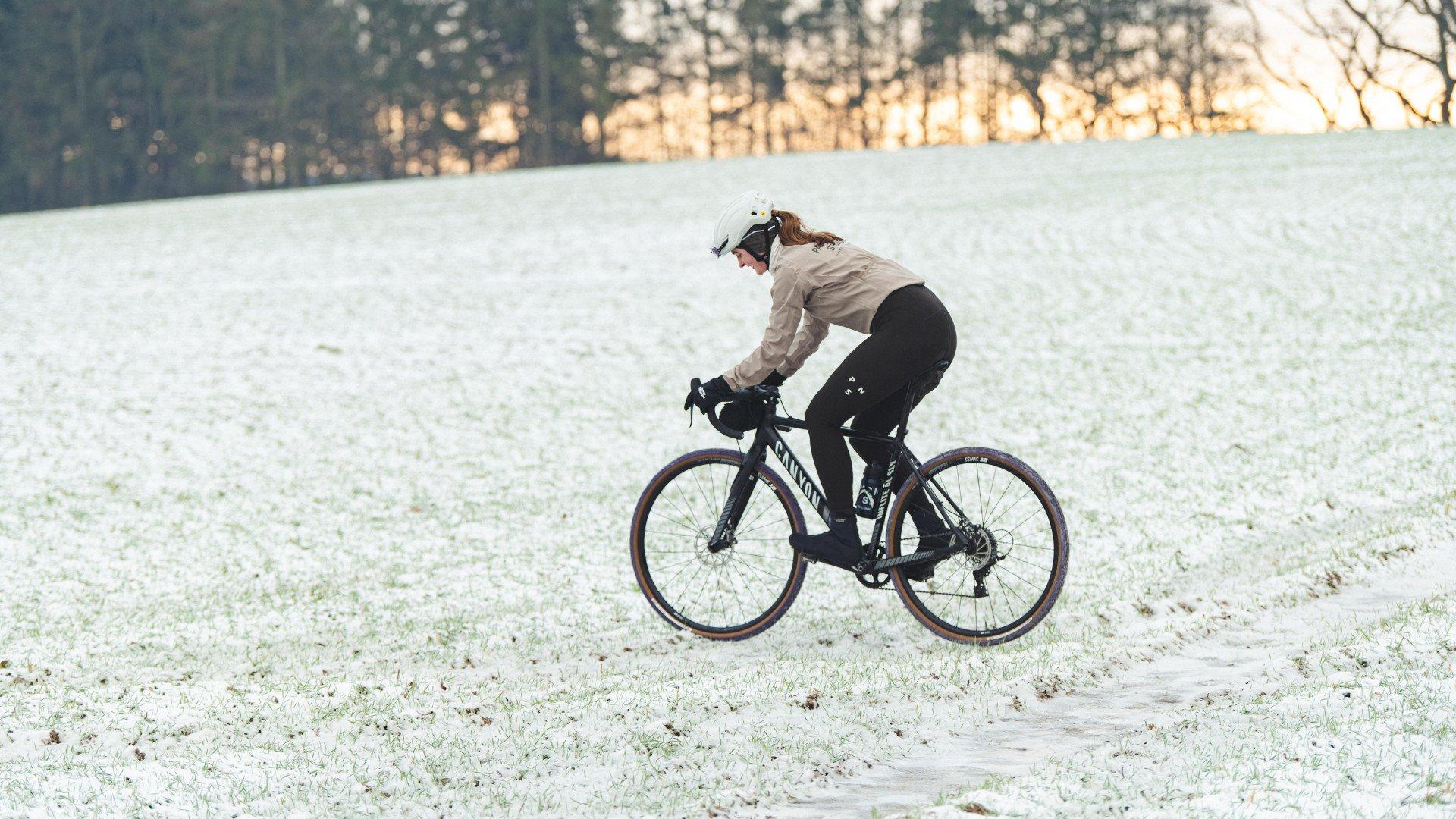 Model wearing beige jacket and black tights cycling on frost-covered field in winter landscape
