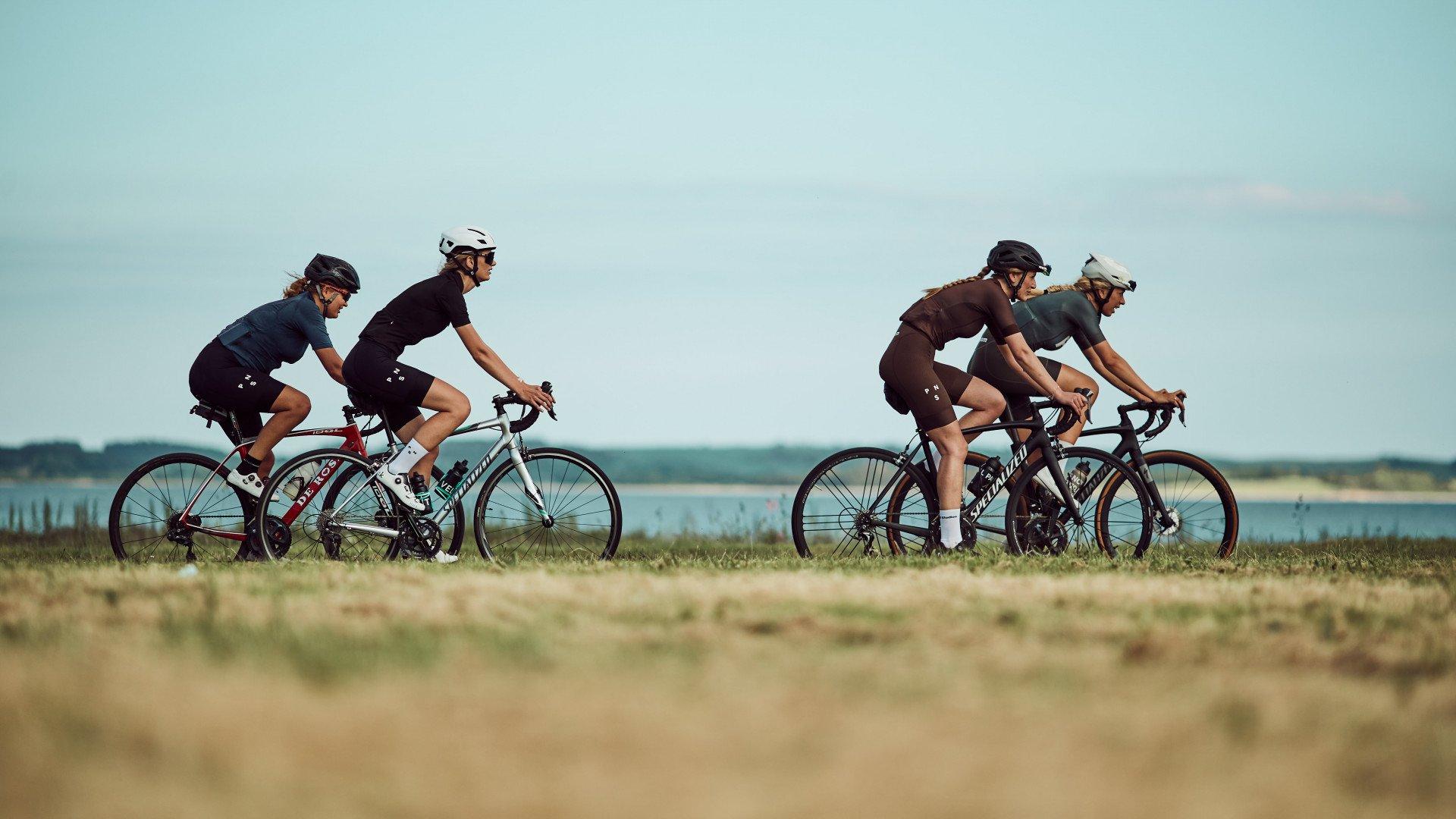 Four cyclists in aerodynamic position riding along coastal road with water and sky in background