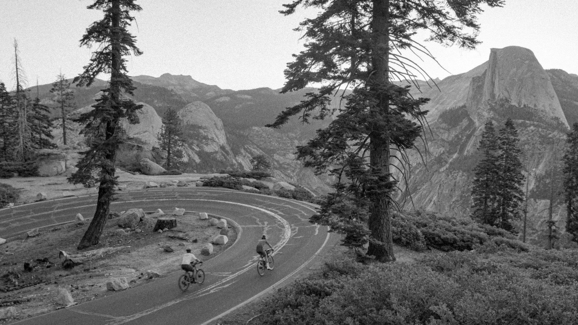 Cyclists on winding mountain road through dramatic landscape with granite cliffs and pine trees