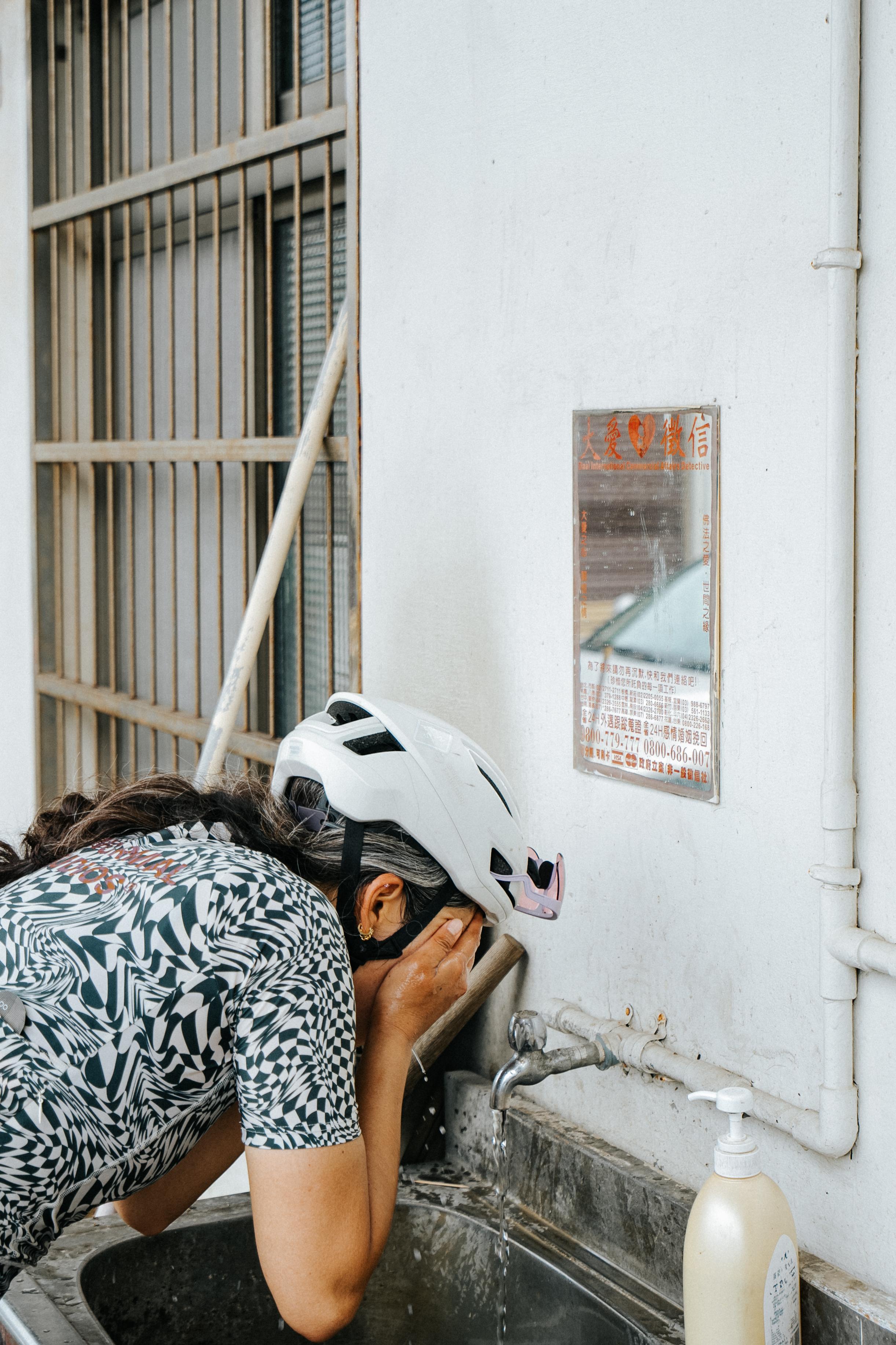 Model wearing white cycling helmet washing face at outdoor water station in black and white patterned jersey