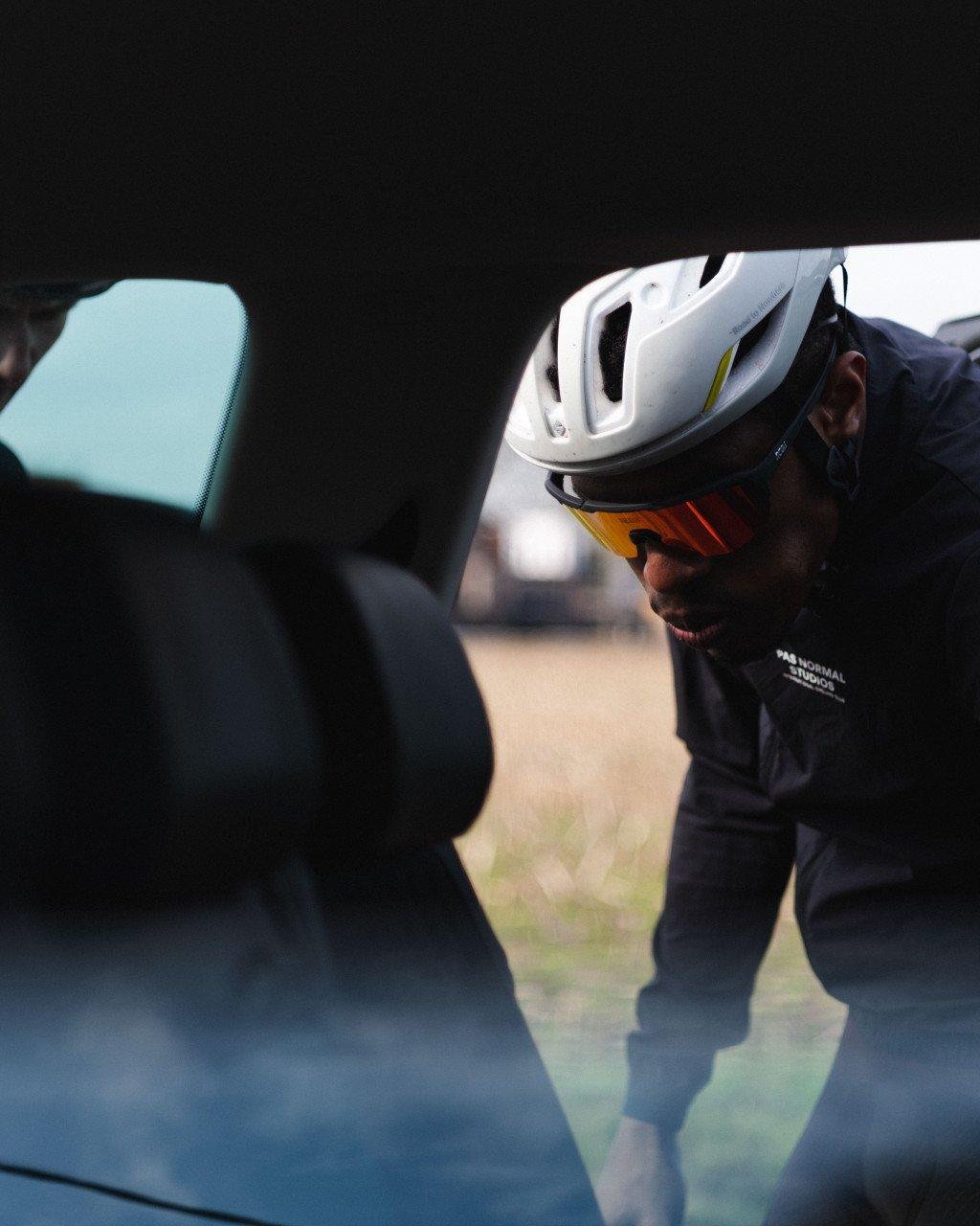 Model wearing dark jersey with logo detail, white helmet and orange sunglasses viewed through car window