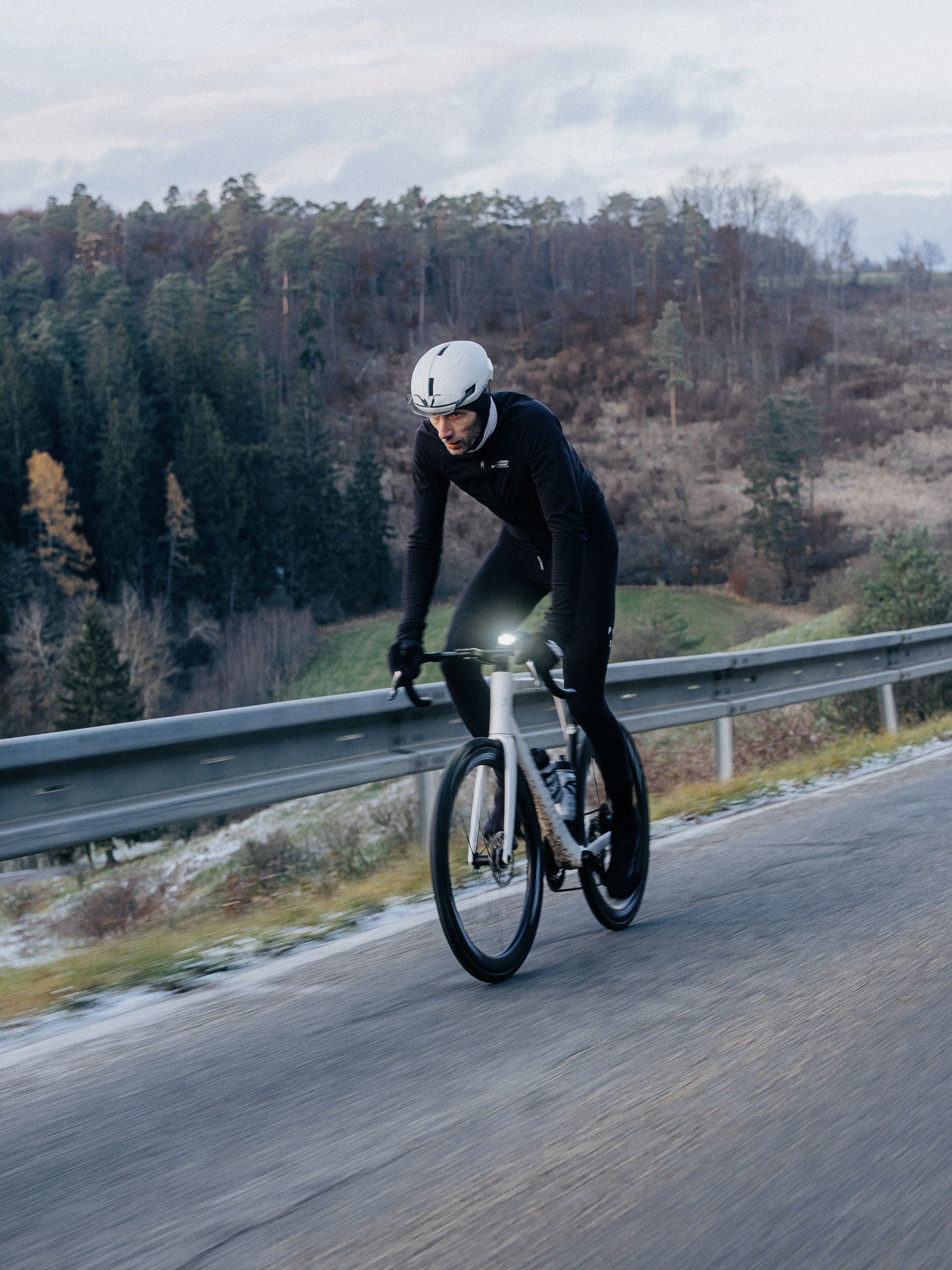 Model wearing black cycling kit on time trial bike with white helmet on road through hilly landscape